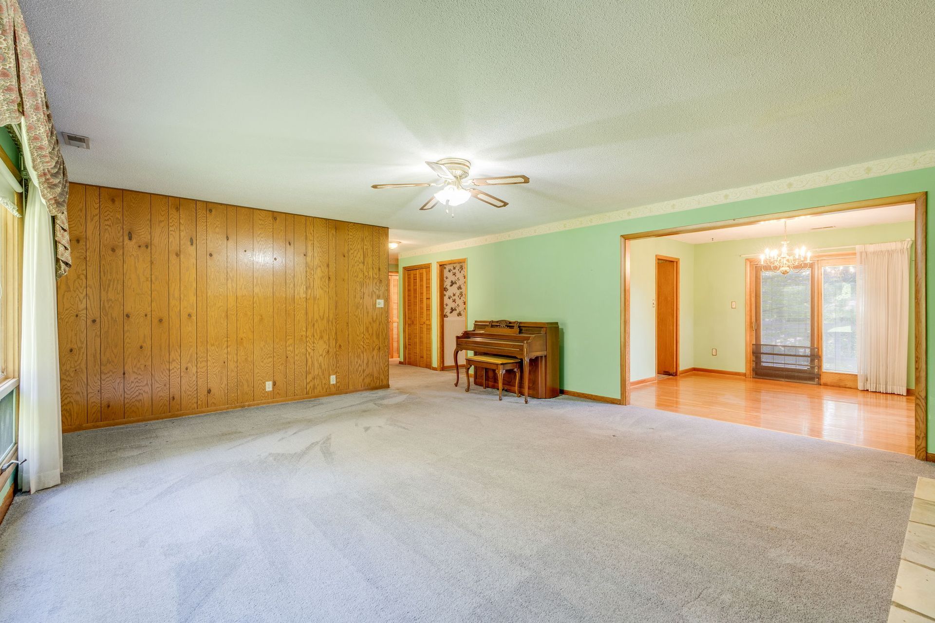 Empty living room with carpet, wood-paneled wall, ceiling fan, and open doorway to hardwood dining area