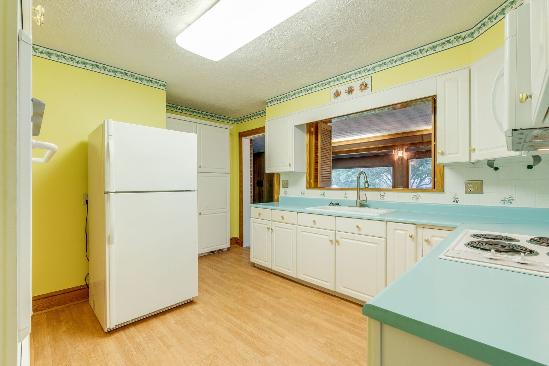 Bright yellow kitchen with white appliances, mint countertops, and a large window over the sink.
