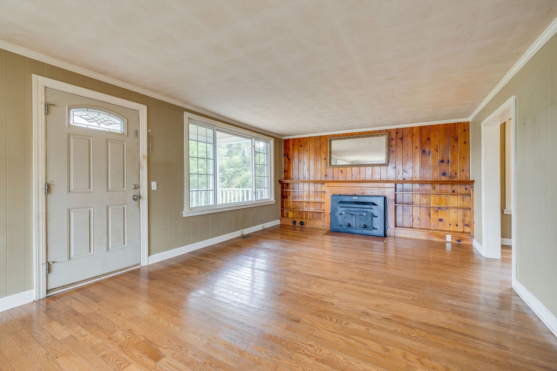 Empty living room with hardwood floors, beige walls, large window, and wood-paneled fireplace wall.