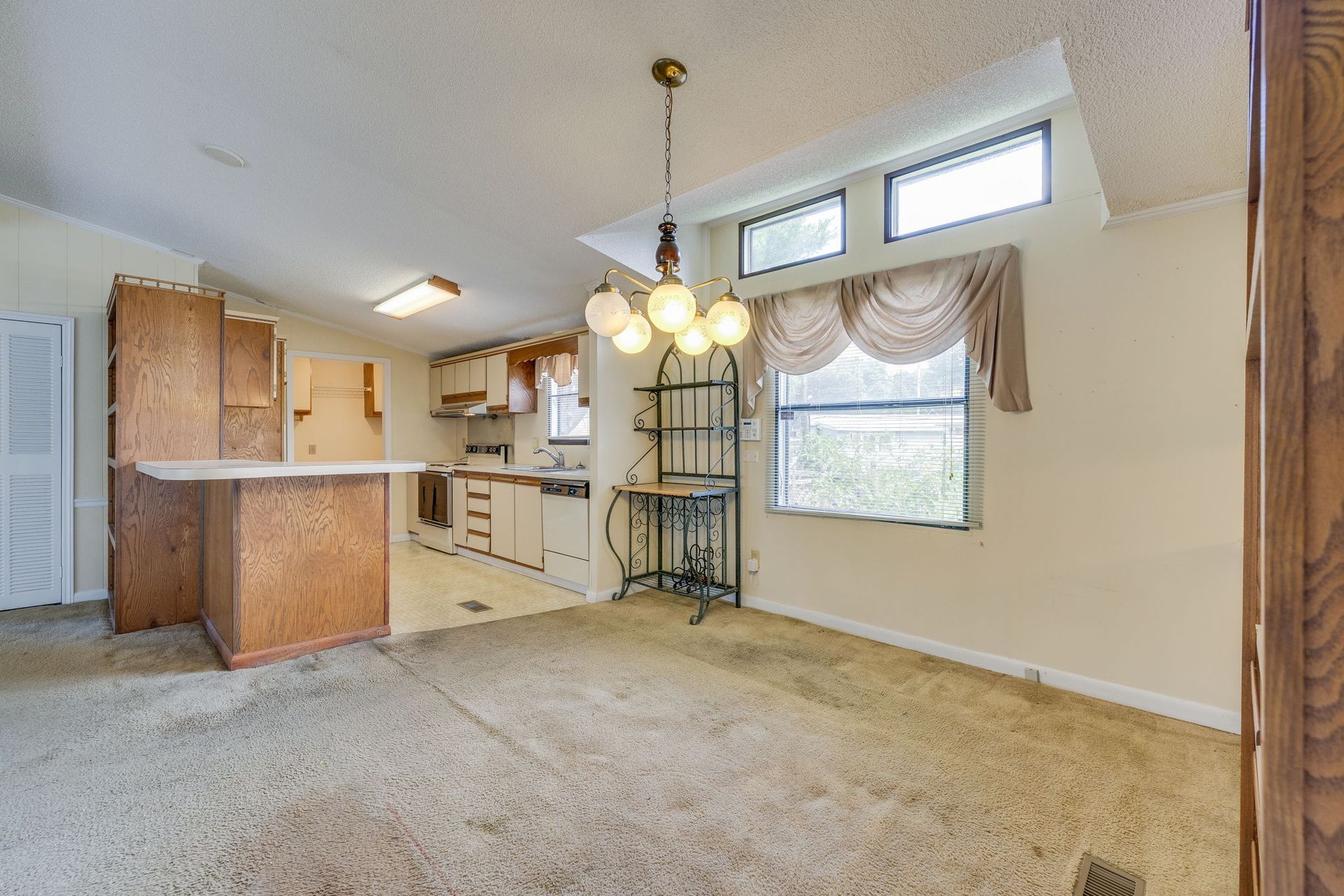Empty beige living room with carpet, kitchen bar, chandelier, and high windows