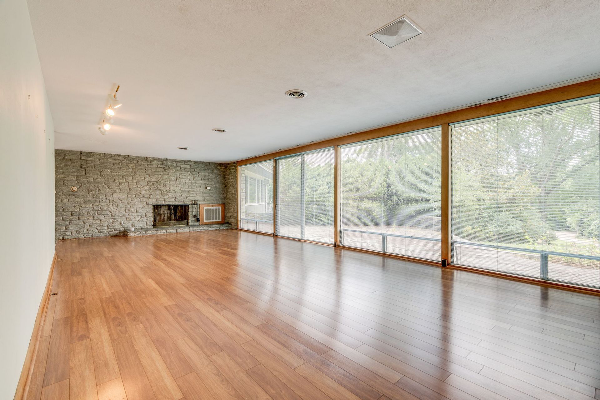Empty modern living room with wood floors, stone wall, and floor-to-ceiling windows overlooking trees