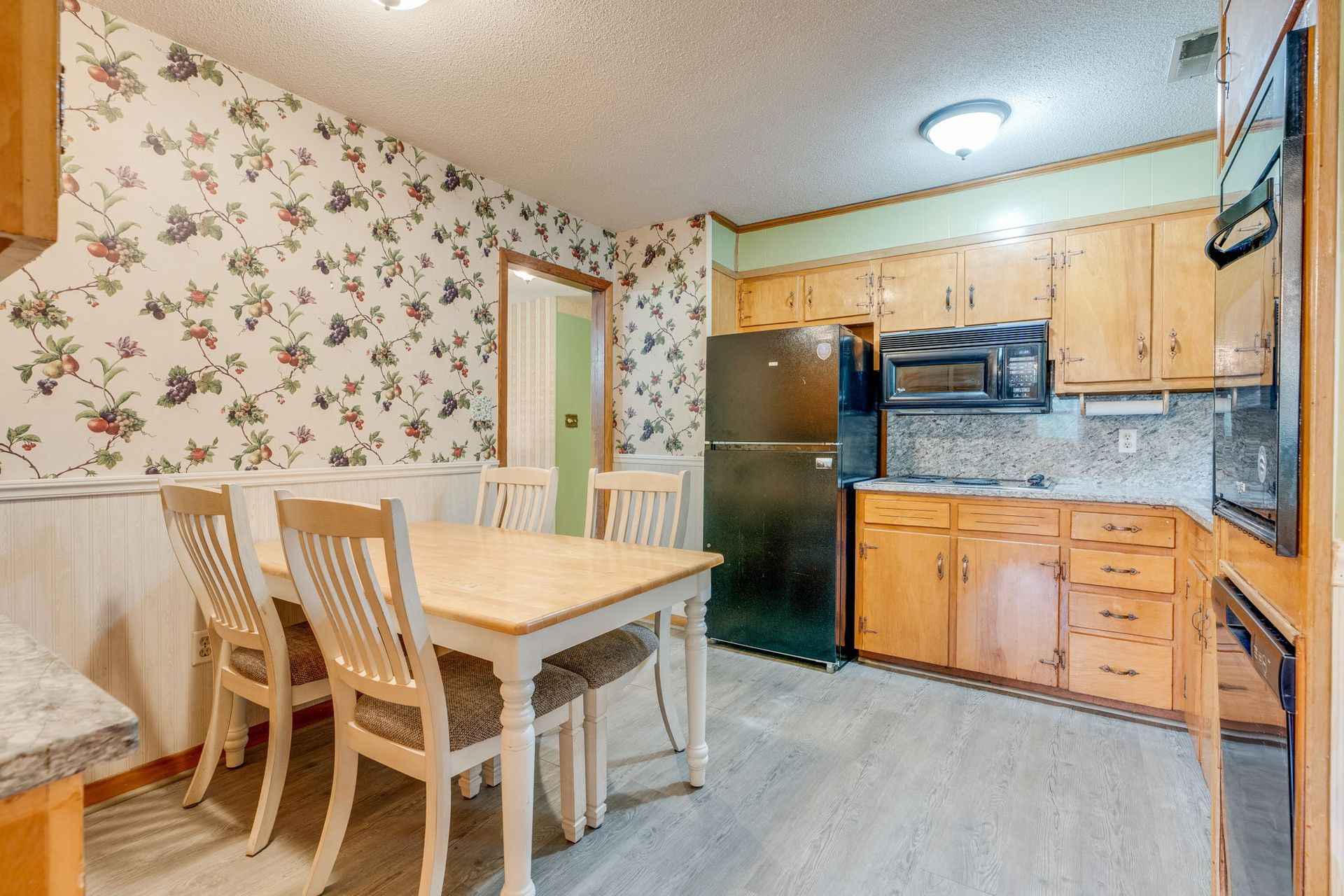 Dining room and kitchen with floral wallpaper, wooden table and chairs, black refrigerator, and light wood cabinets.
