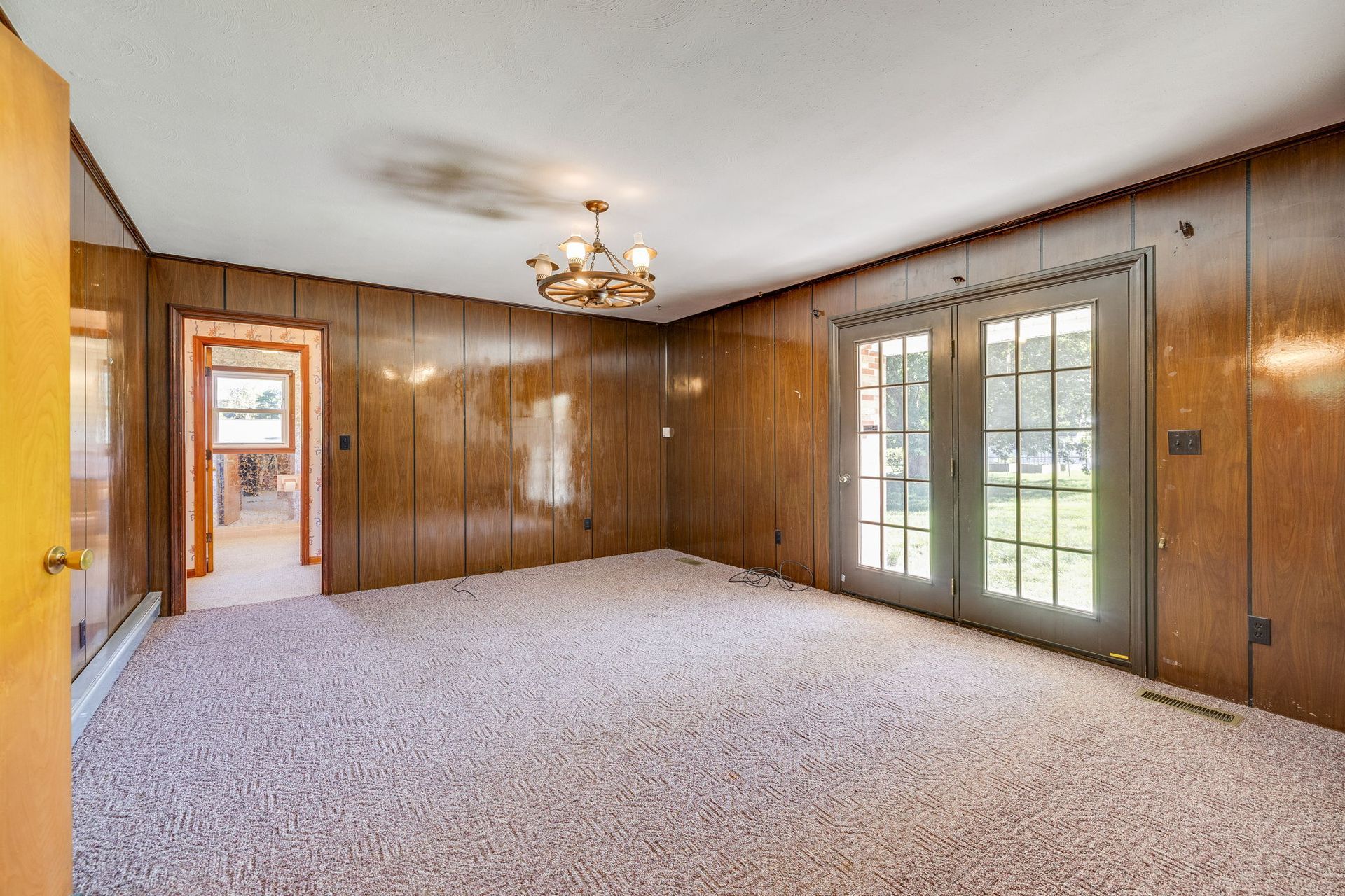 Empty wood-paneled living room with carpet, chandelier, and French doors to a yard.