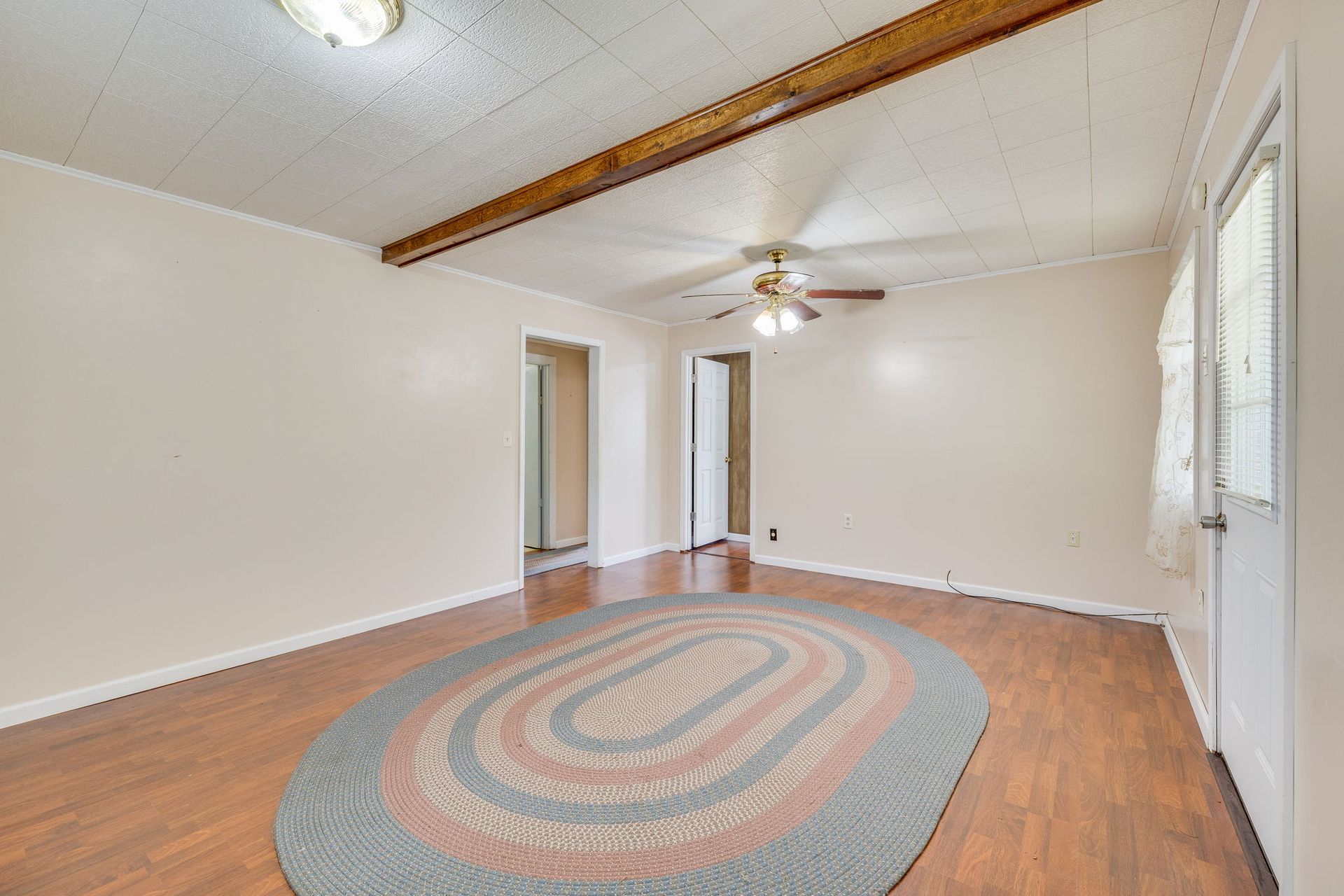 Empty living room with hardwood floors, white walls, exposed beam, ceiling fan, and oval rug.