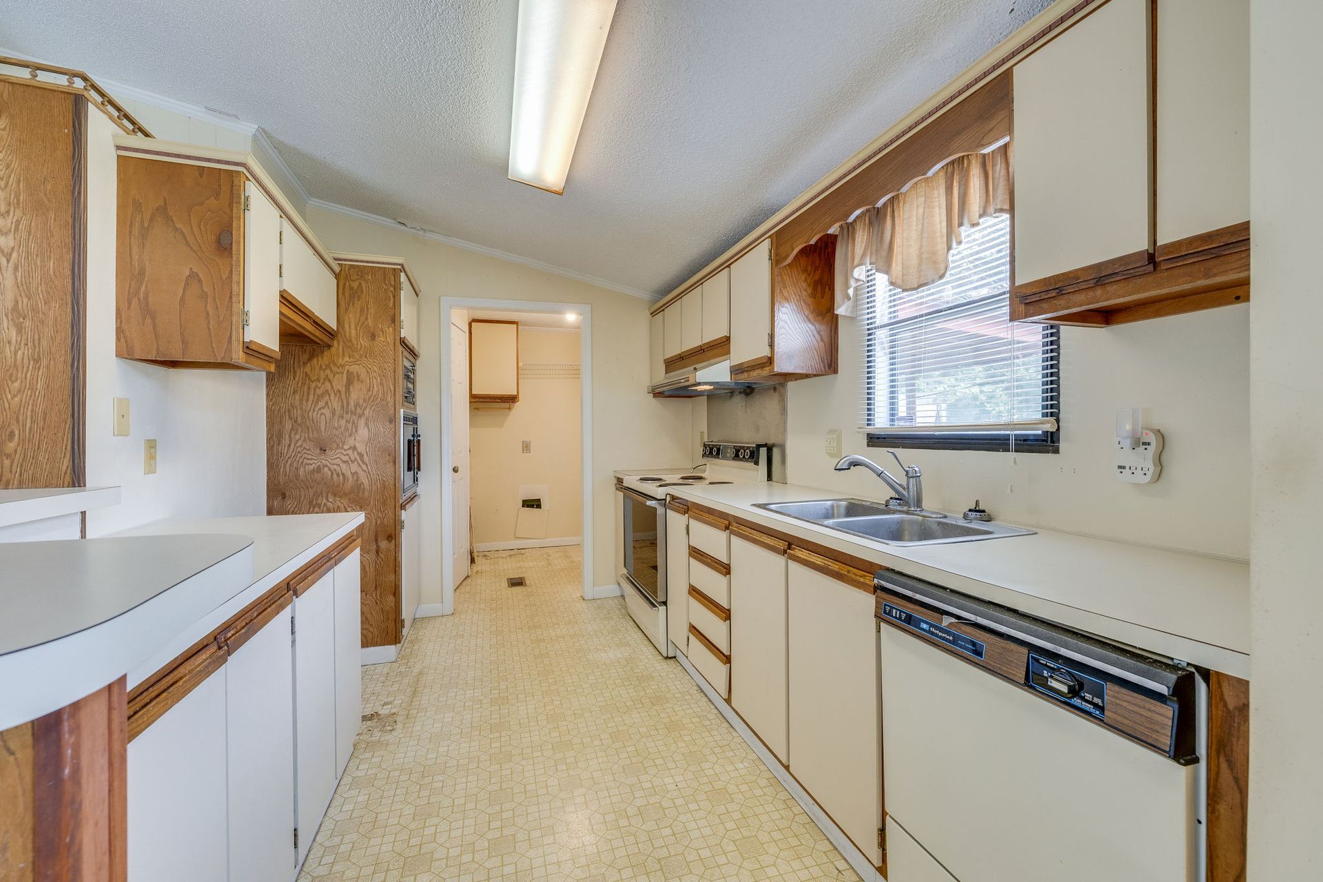 Narrow galley kitchen with wood cabinets, beige counters, white appliances, and a window over the sink