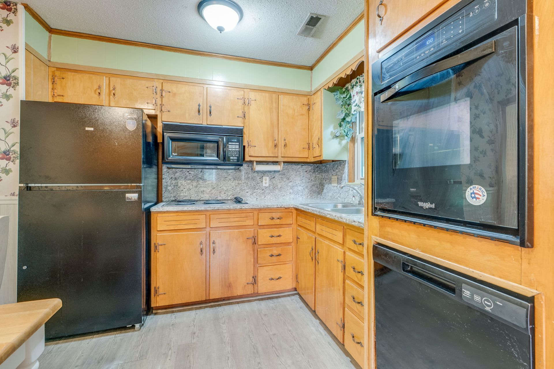 Compact kitchen with black appliances, light wood cabinets, and tiled backsplash under a ceiling light