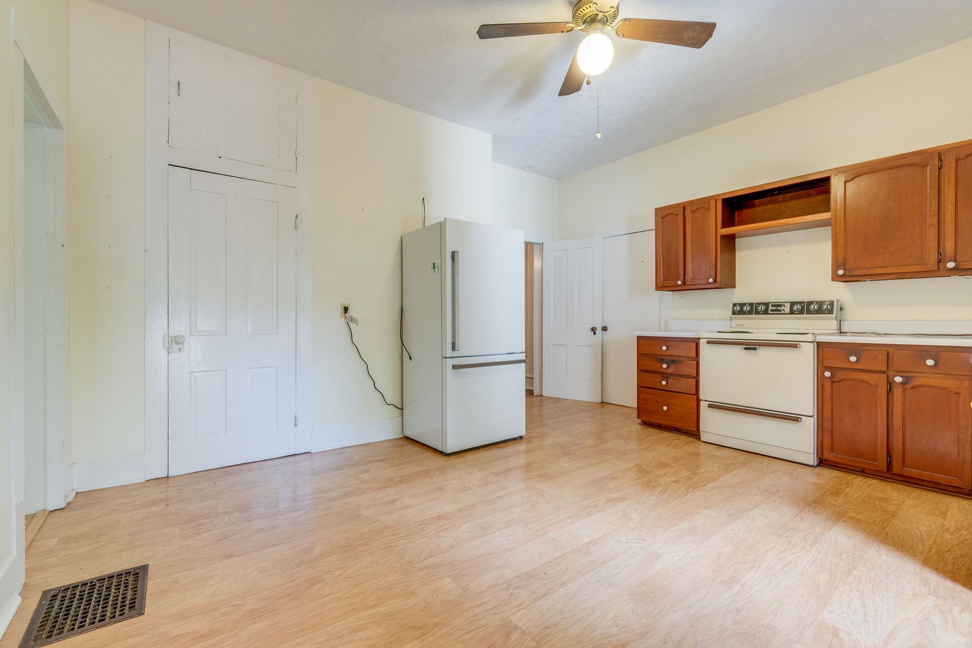 Empty kitchen with white appliances, wood cabinets, beige carpet, and a ceiling fan on.