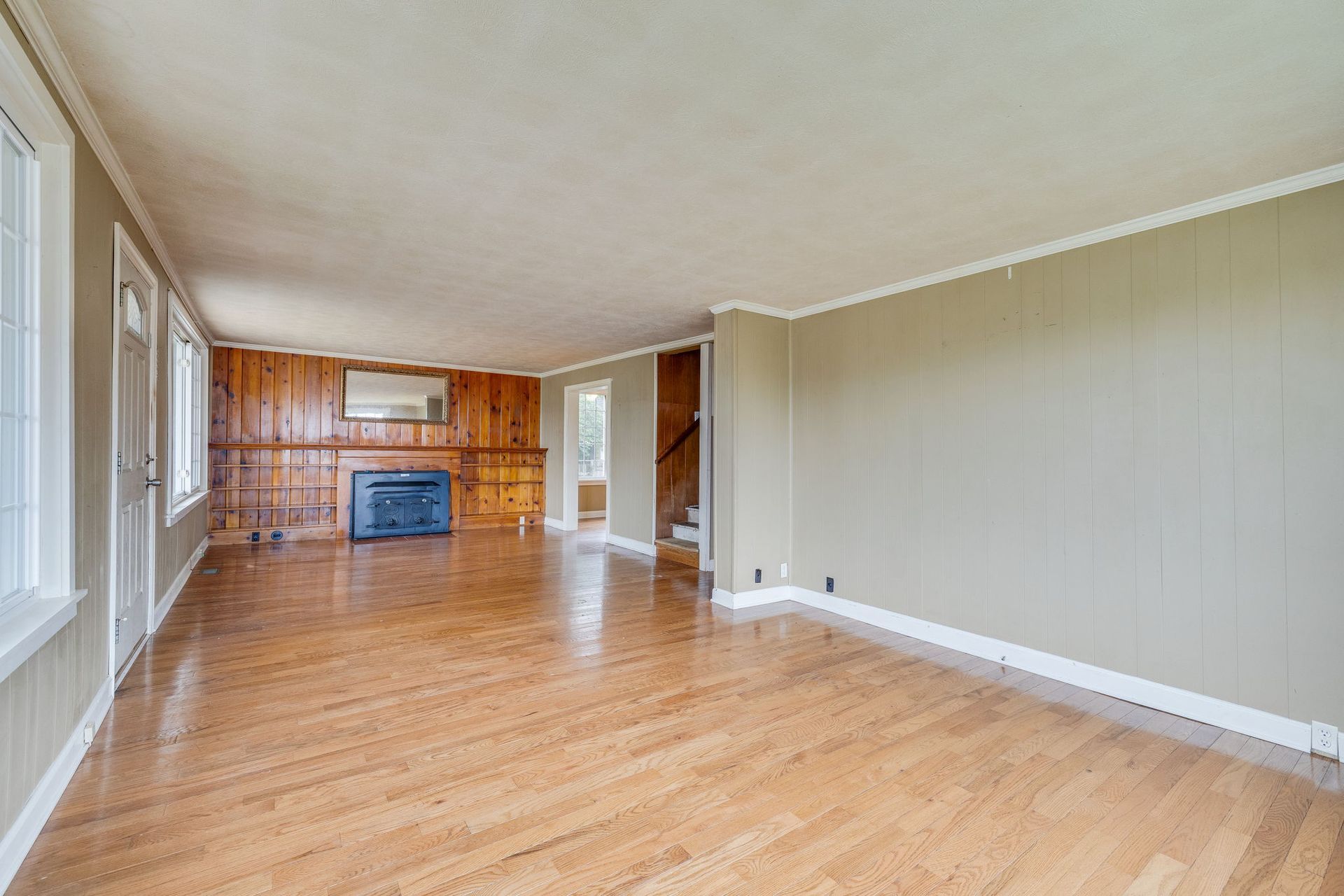 Empty living room with hardwood floors, beige walls, and a brick fireplace at the far end.