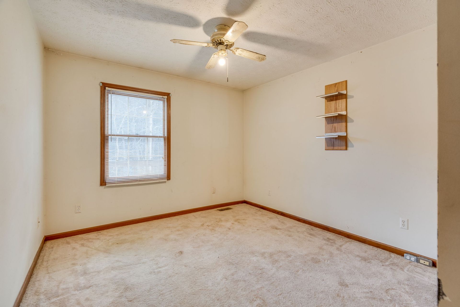 Empty room with beige walls, window with blinds, ceiling fan, and built-in shelving.