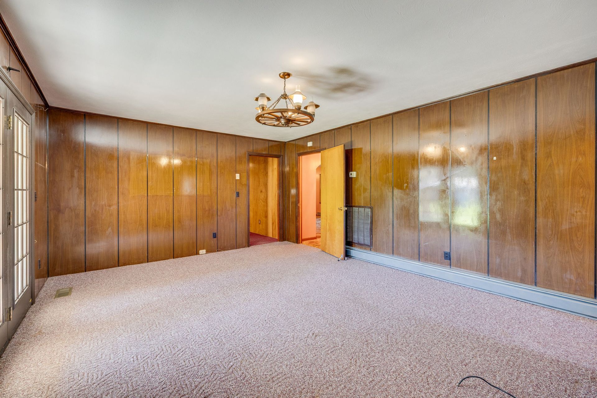 Empty wood-paneled living room with beige carpet, chandelier, and doorway to a lit hall.