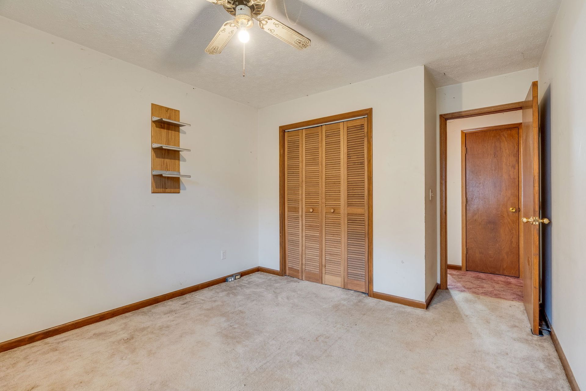 Empty bedroom with beige carpet, wooden closet doors, and open doorway. Ceiling fan and small shelves on wall.