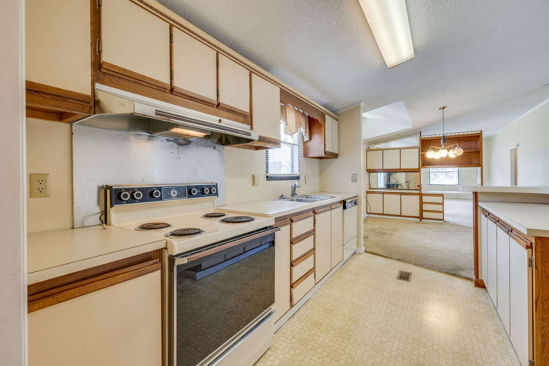 Empty beige kitchen with white cabinets, brown countertops, and a view into a bright adjoining room