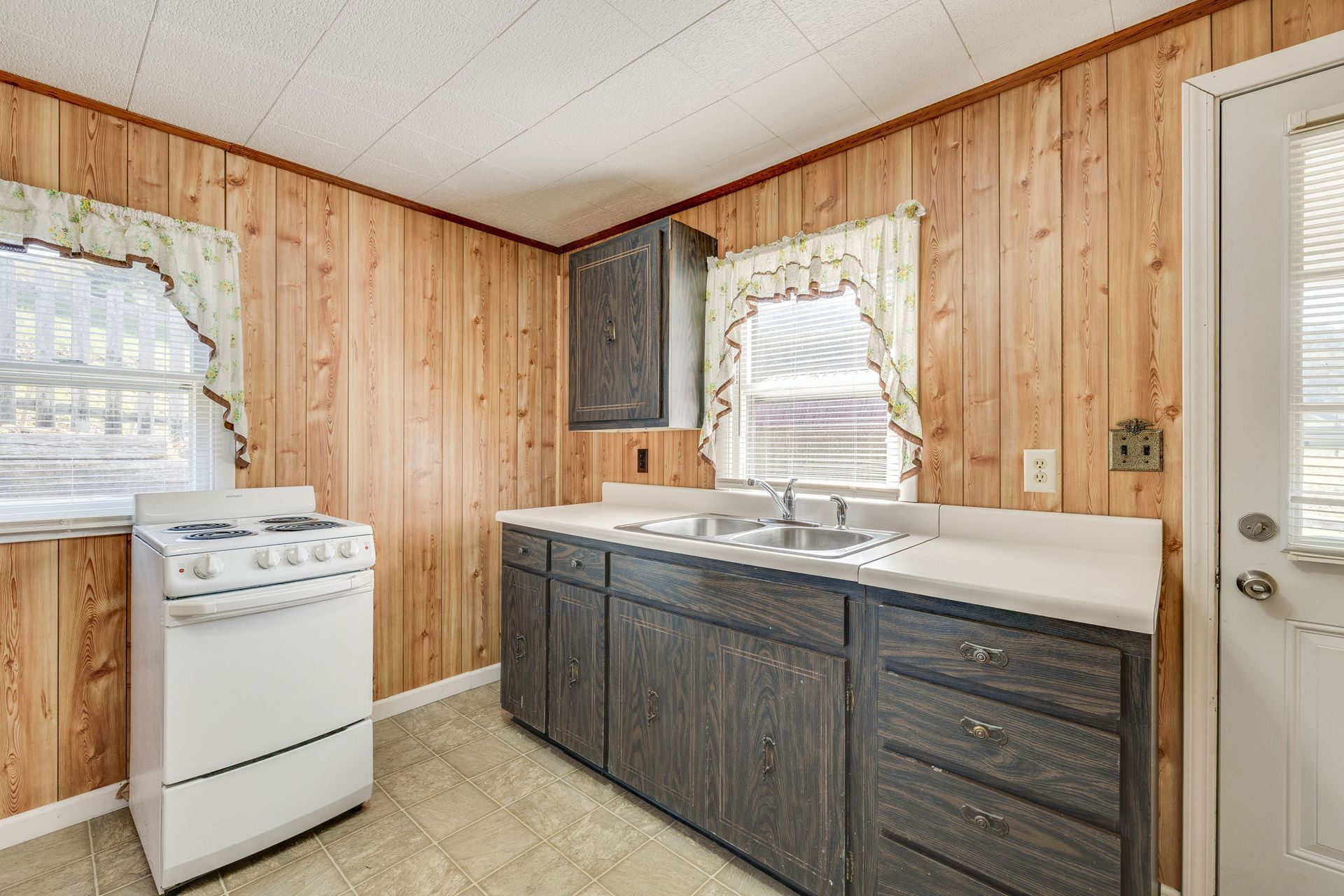 Small rustic kitchen with wood paneling, white stove, and dark cabinets under two windows
