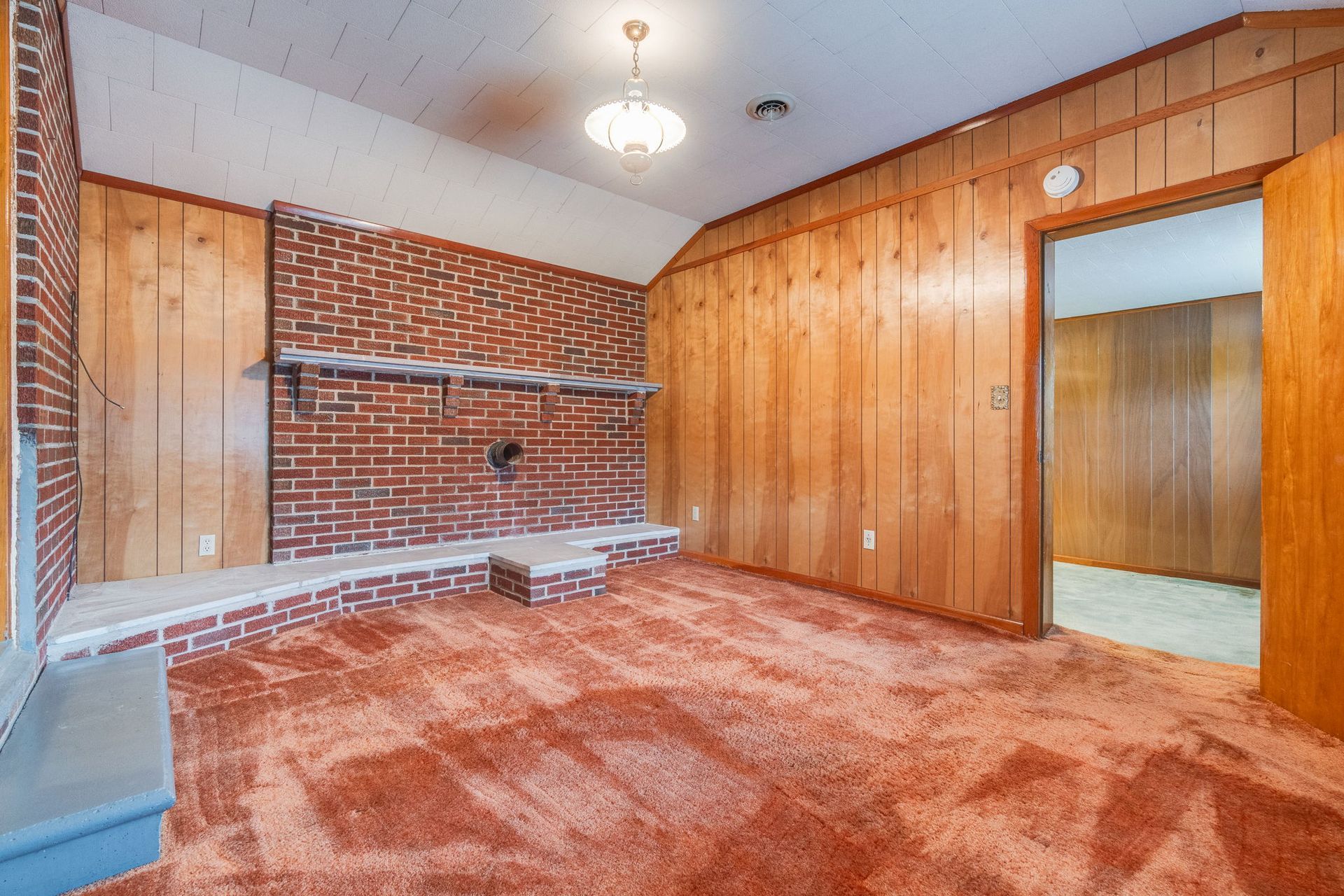 Empty retro bedroom with wood-paneled walls, brick fireplace, and orange carpet