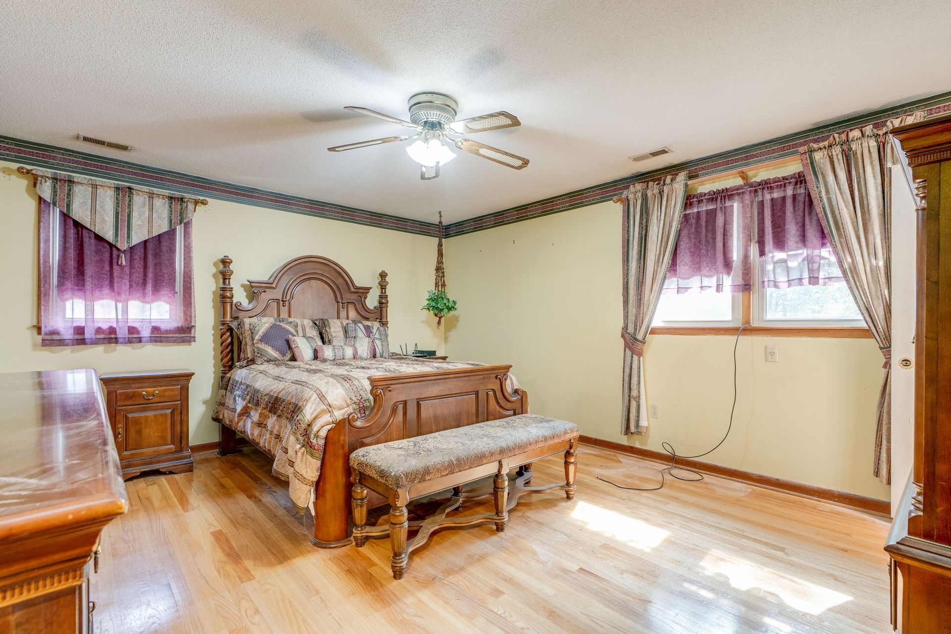 Spacious bedroom with a wooden bed, bench, purple curtains, and ceiling fan in soft daylight