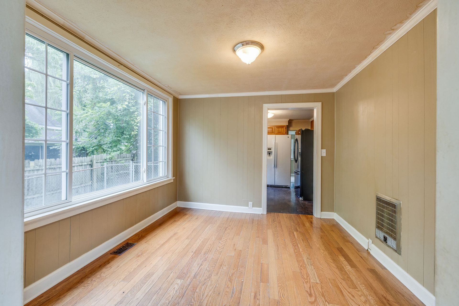 Empty beige living room with hardwood floors, large window, and doorway to a kitchen area