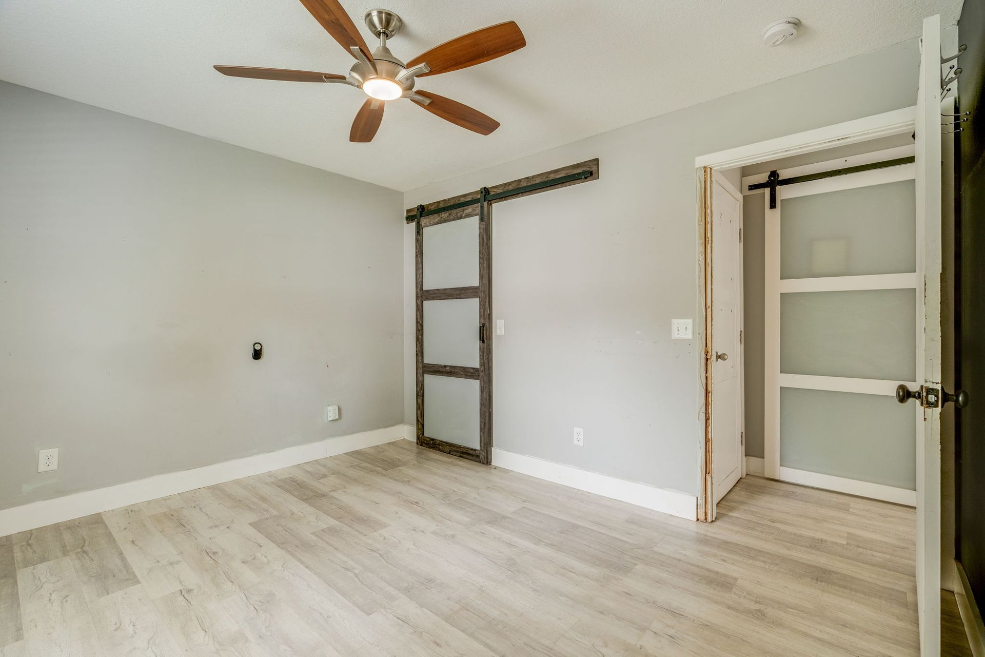 Empty bedroom with light gray walls, wood-look flooring, ceiling fan, and open closet doors.