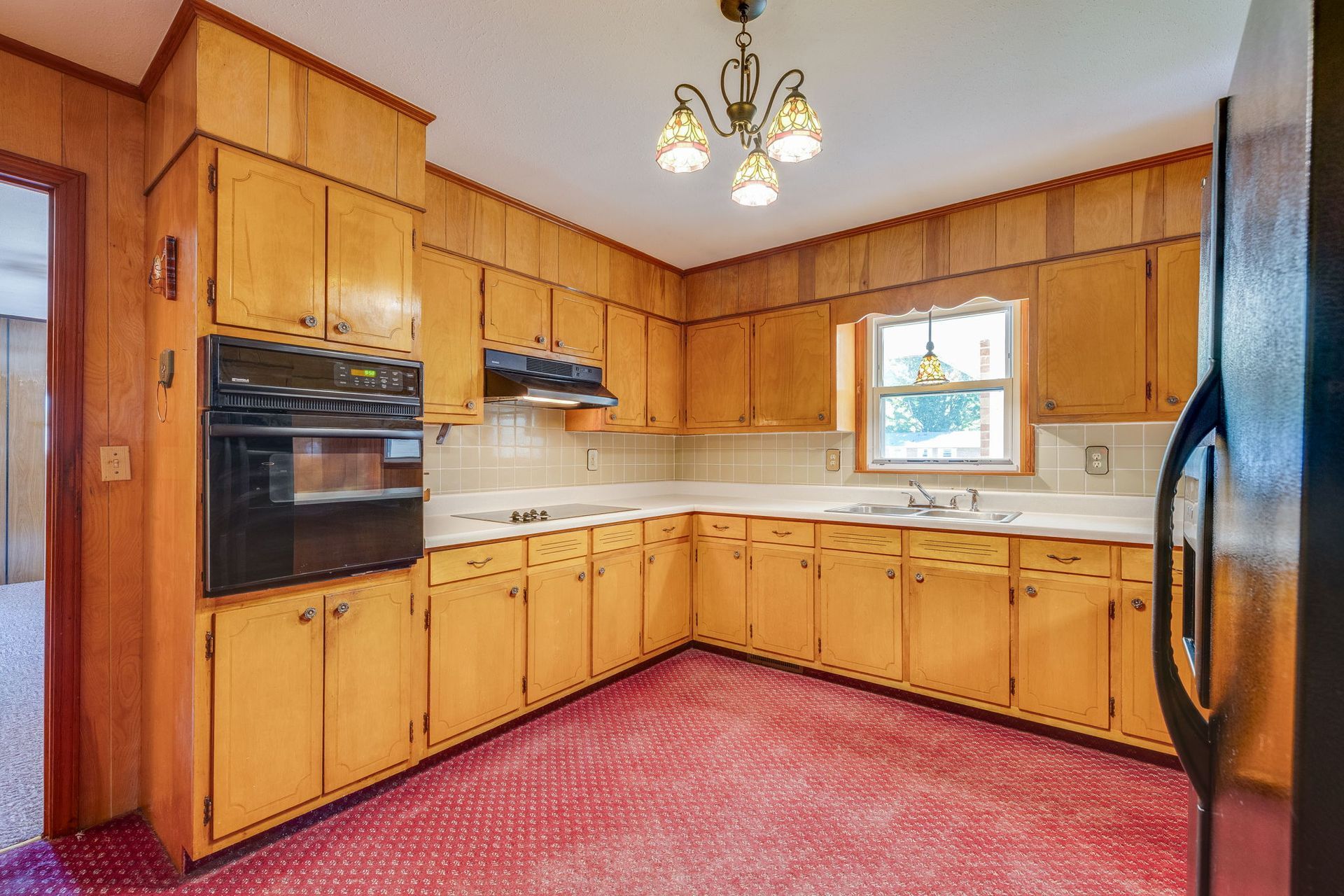 Wooden kitchen with U-shaped cabinets, black appliances, red carpet, and a window over the sink