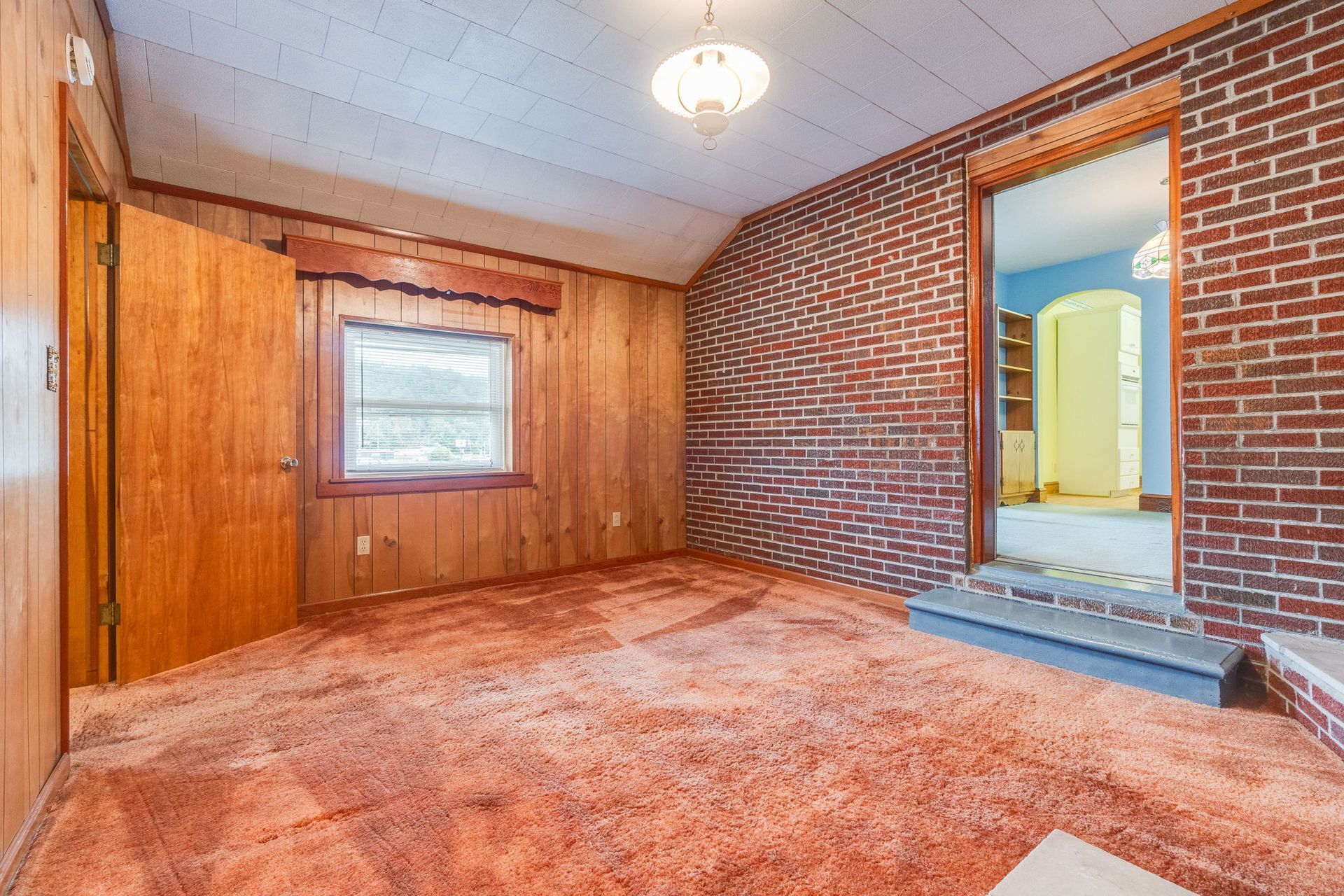 Empty wood-paneled room with red carpet, brick walls, and an open doorway to a lit hallway