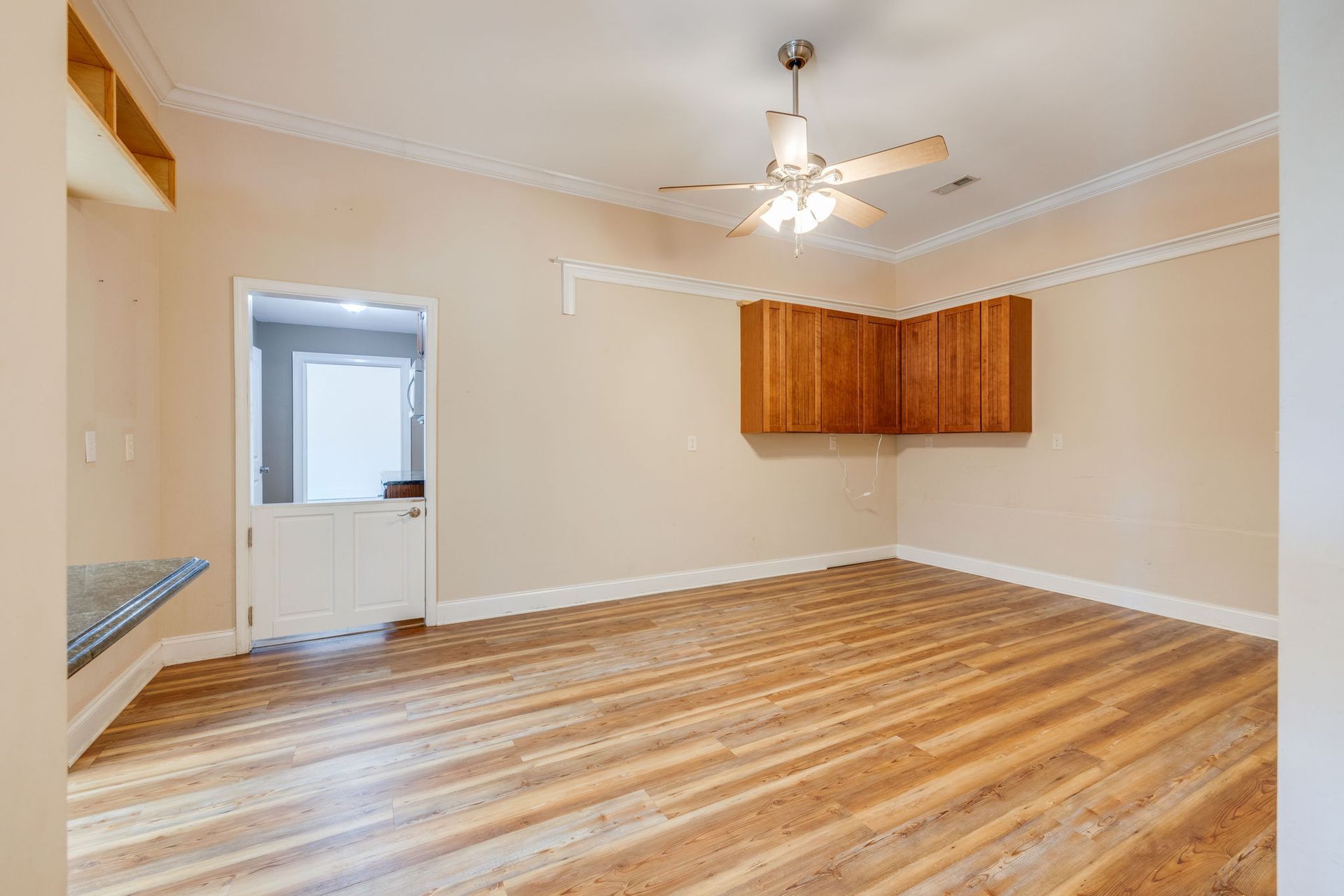 Empty beige room with hardwood floors, ceiling fan, and wooden wall cabinets.