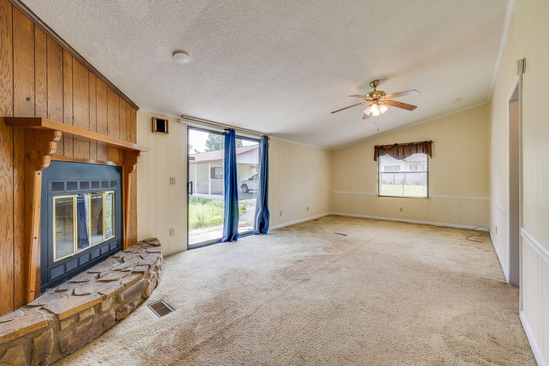 Empty beige living room with fireplace, ceiling fan, carpet, and sliding glass doors to backyard.