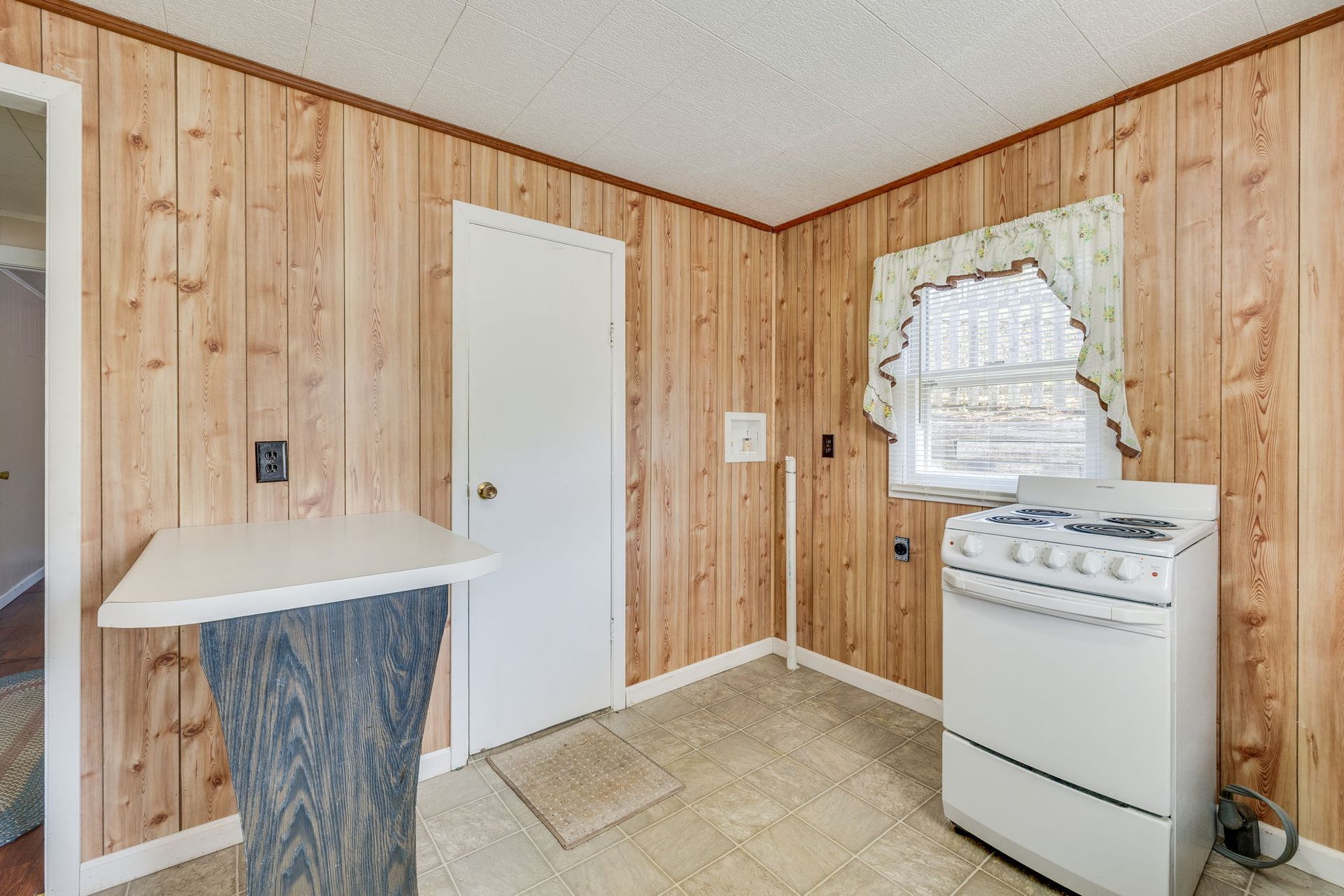 Small kitchen with wood-paneled walls, white stove, and a window with lace curtains