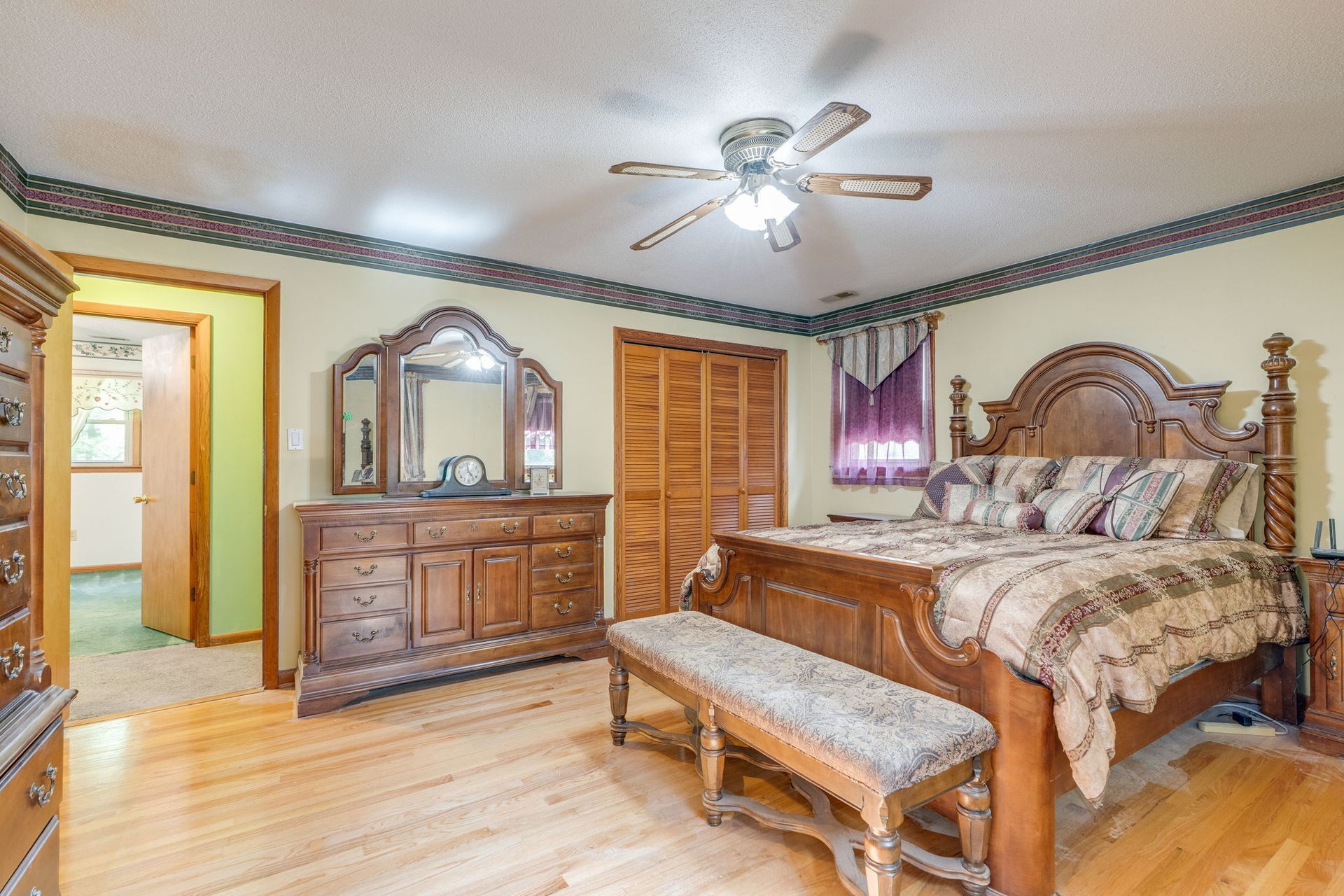 Spacious bedroom with wooden furniture, carved bed, dresser, bench, and ceiling fan, in warm neutral tones