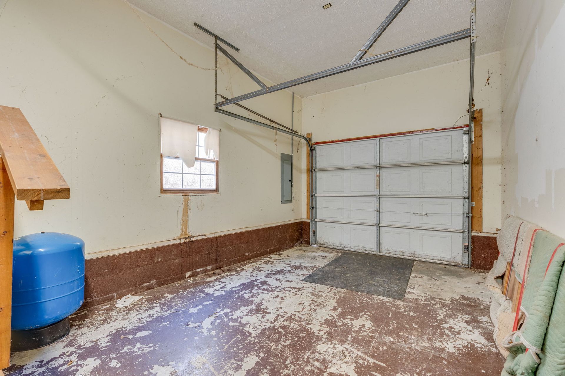 Interior of a garage with peeling paint, a closed garage door, window, and a blue water tank.