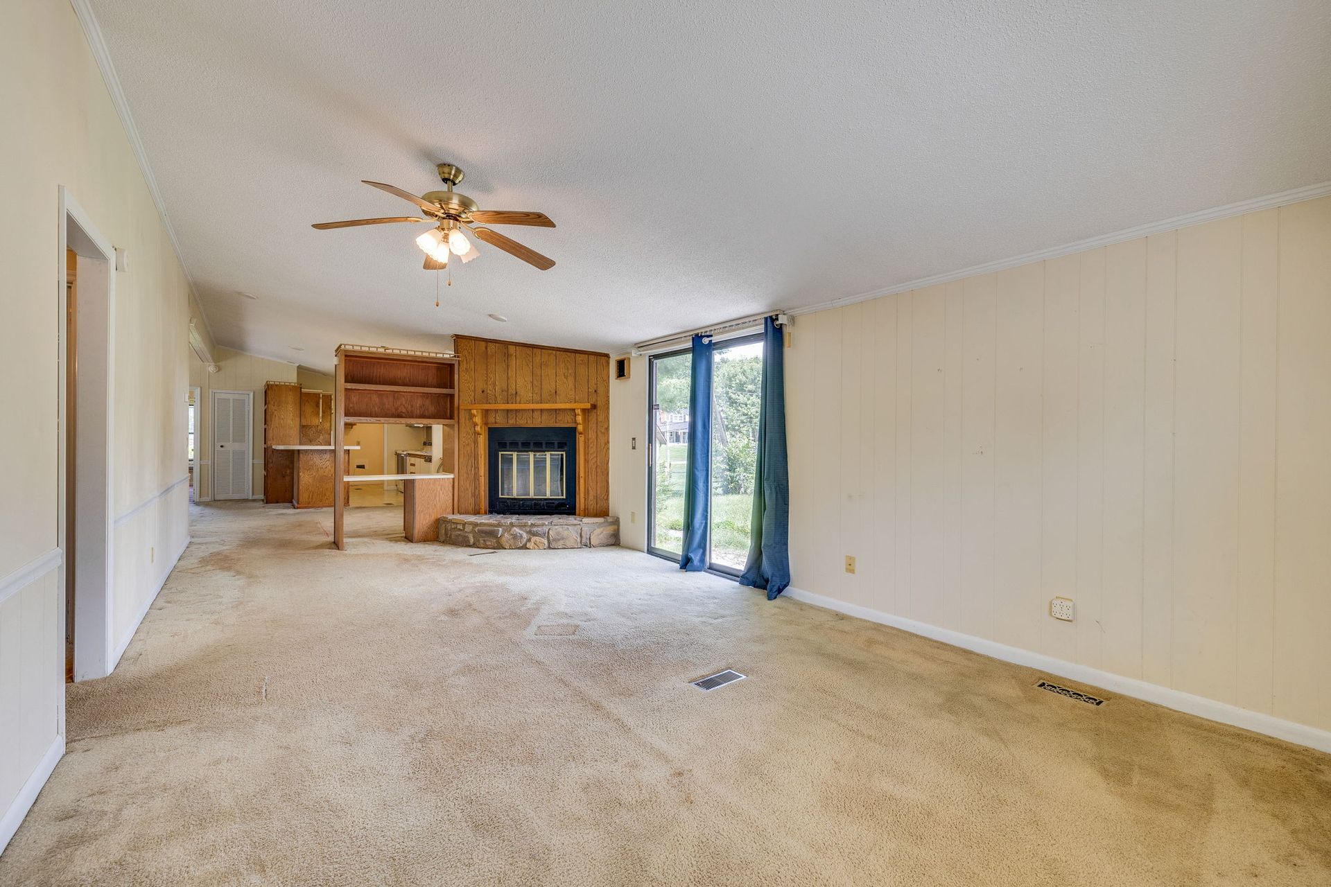 Empty beige living room with carpet, fireplace, ceiling fan, and sliding glass doors.