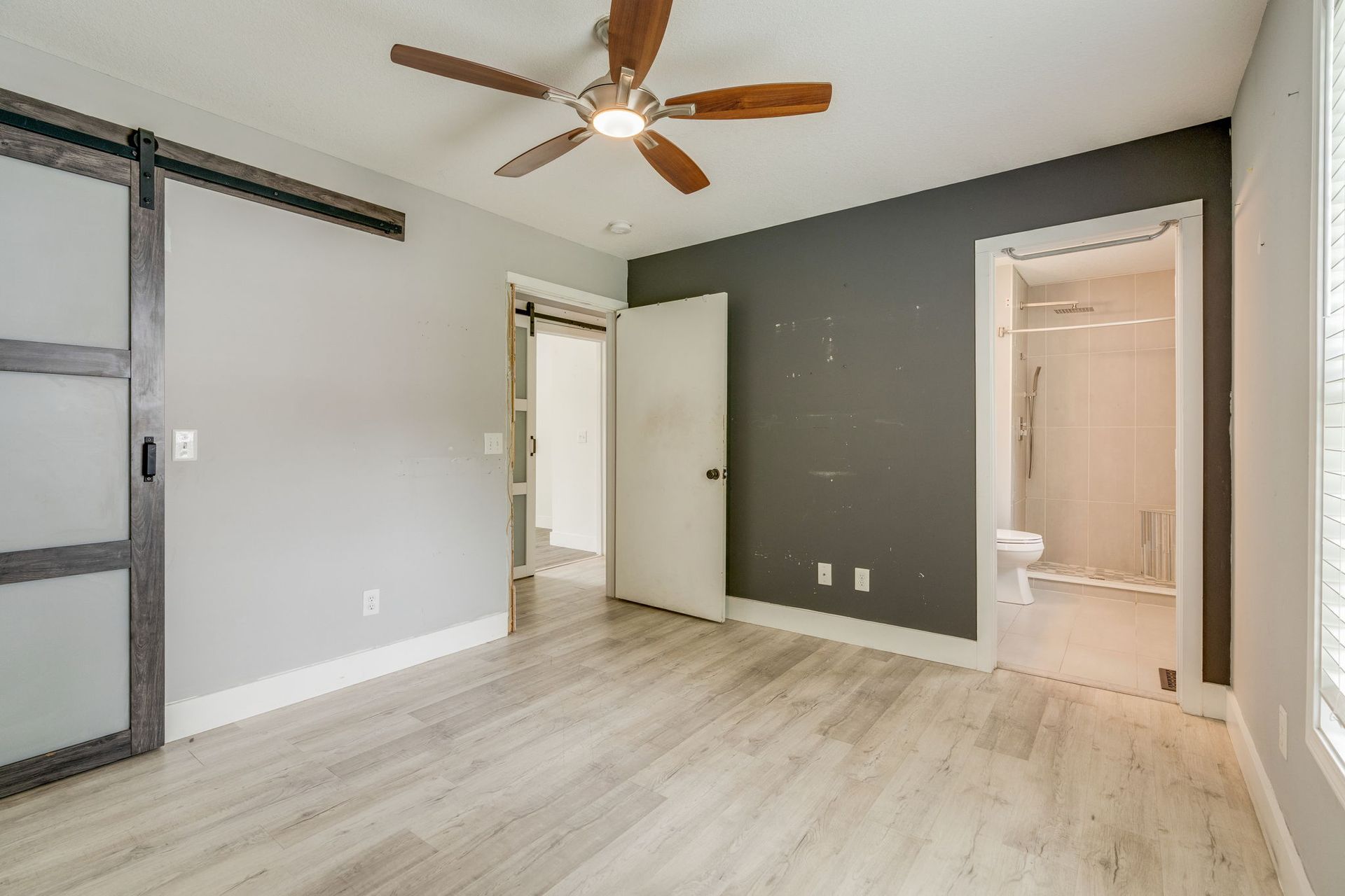 Empty bedroom with light wood floors, gray accent wall, ceiling fan, and open door to bathroom