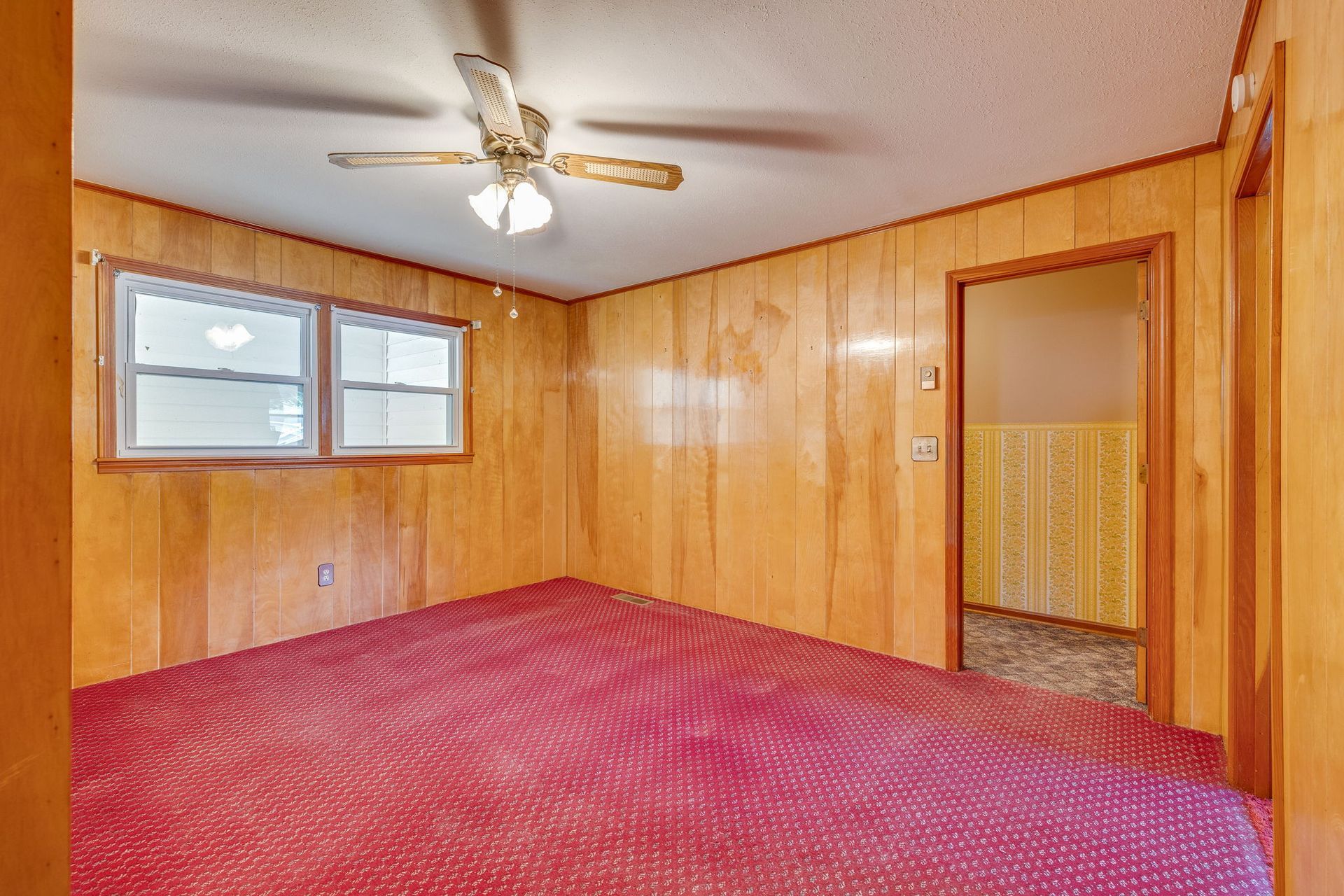 Empty wood-paneled room with red carpet, two windows, ceiling fan, and open doorway