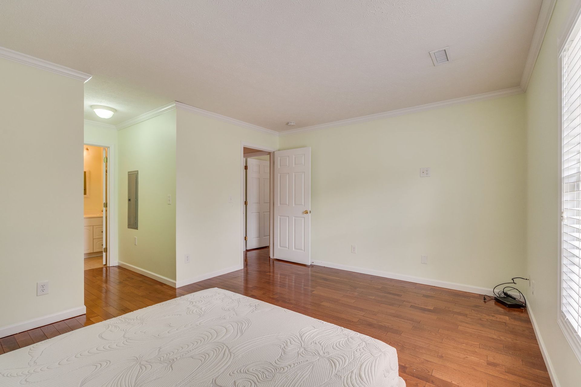 A bedroom with light walls, wood flooring, a white mattress in the foreground, and doorways leading to other rooms.