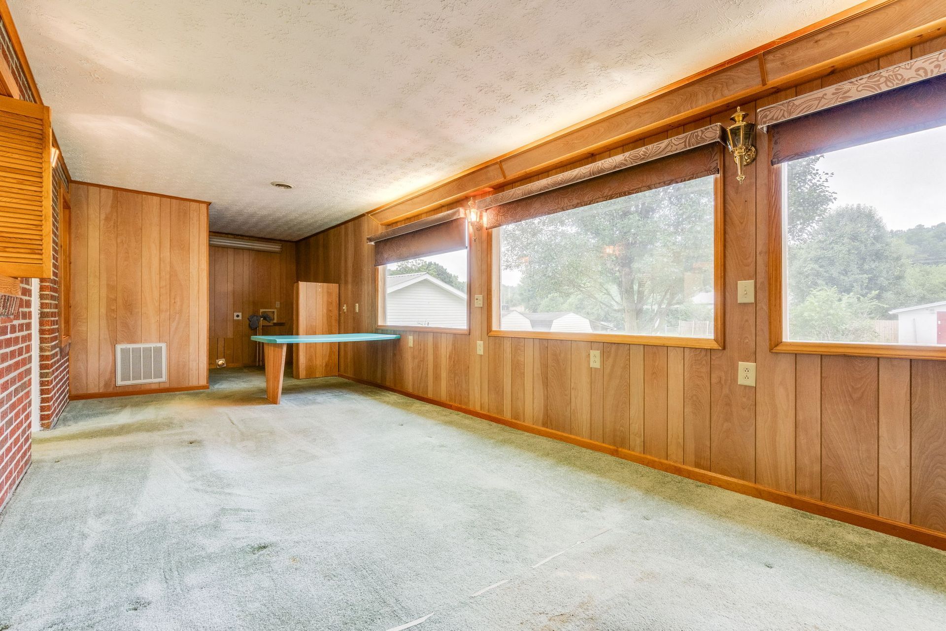 Empty sunroom with wood-paneled walls, green carpet, large windows, and a small table.