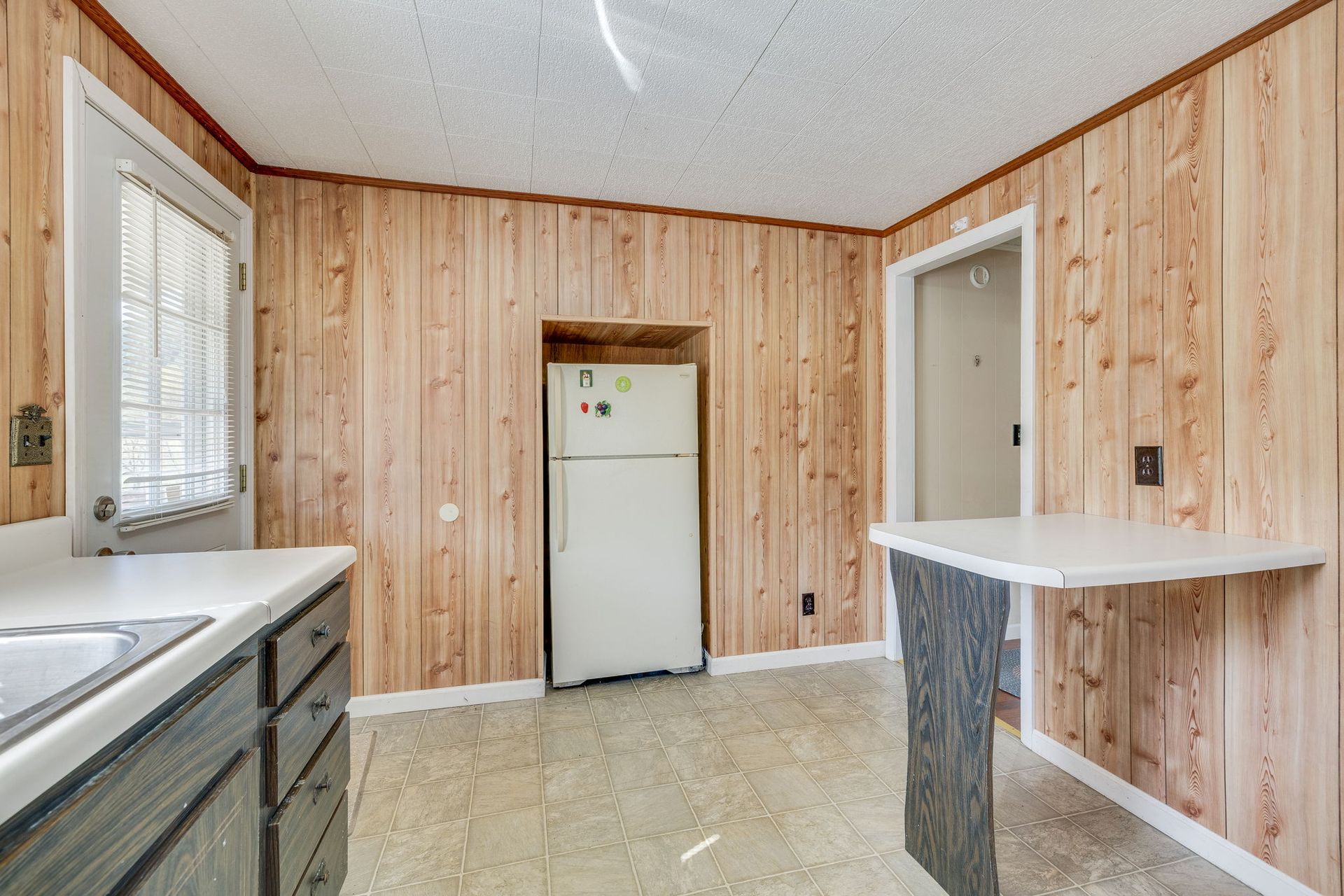 Empty kitchen with wood-paneled walls, white countertops, and a white refrigerator under a window.