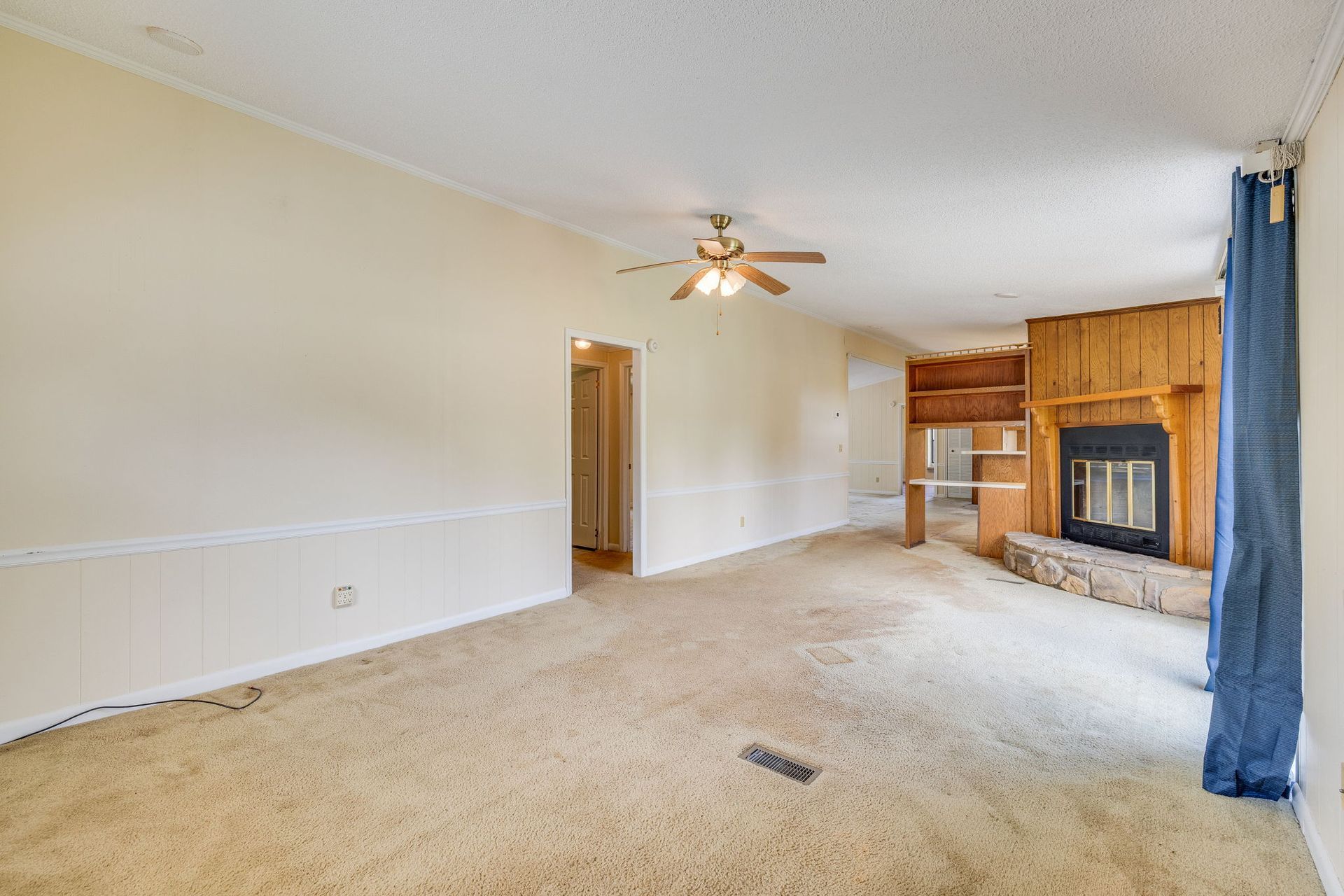 Empty living room with beige carpet, white walls, ceiling fan, and wood fireplace mantel