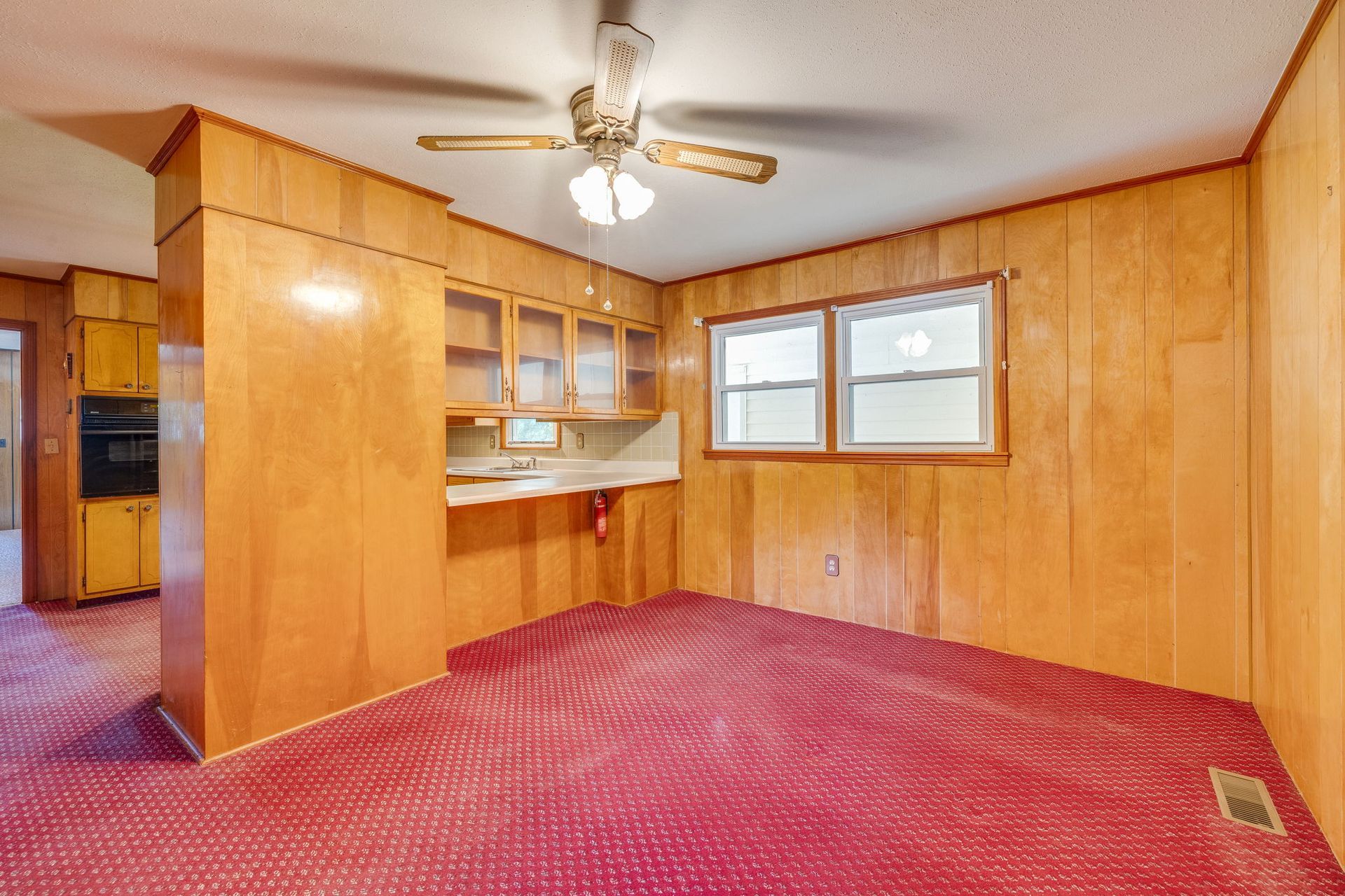 Empty carpeted living room with wood-paneled walls, ceiling fan, pass-through counter, and windows