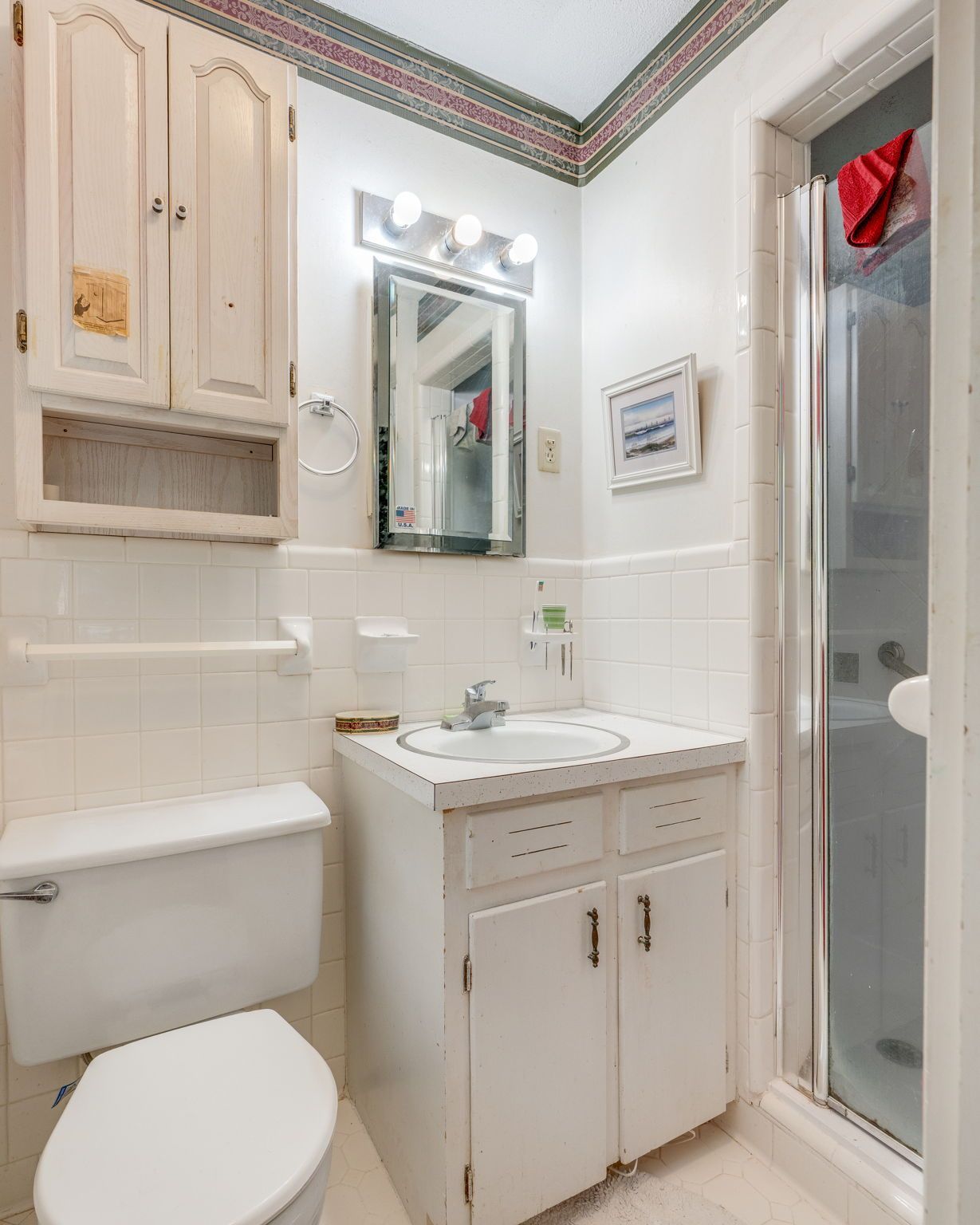 Compact white bathroom with toilet, vanity sink, mirror, wall cabinet, and glass shower door