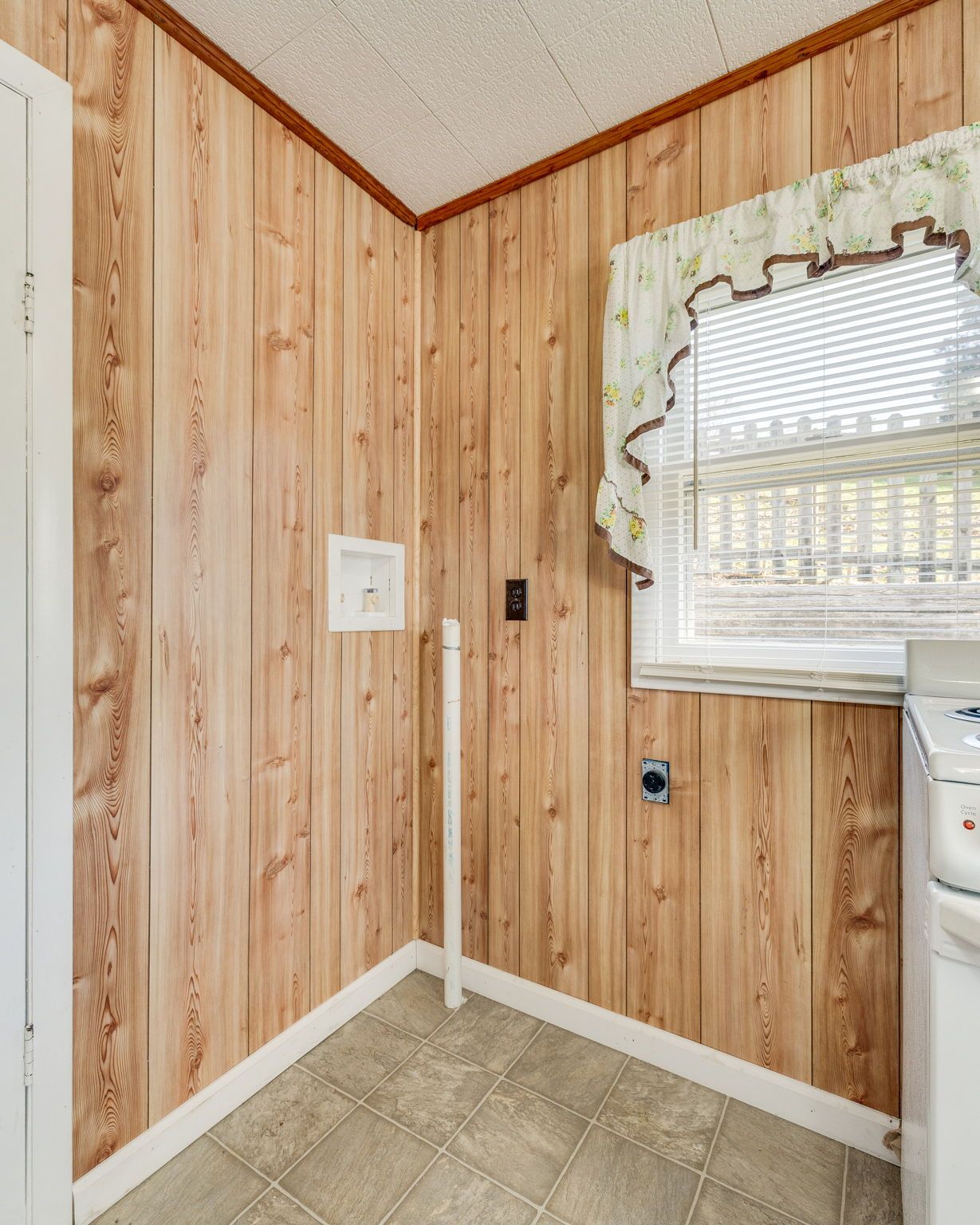 Small laundry room with wood-paneled walls, tiled floor, and a window with a ruffled curtain.