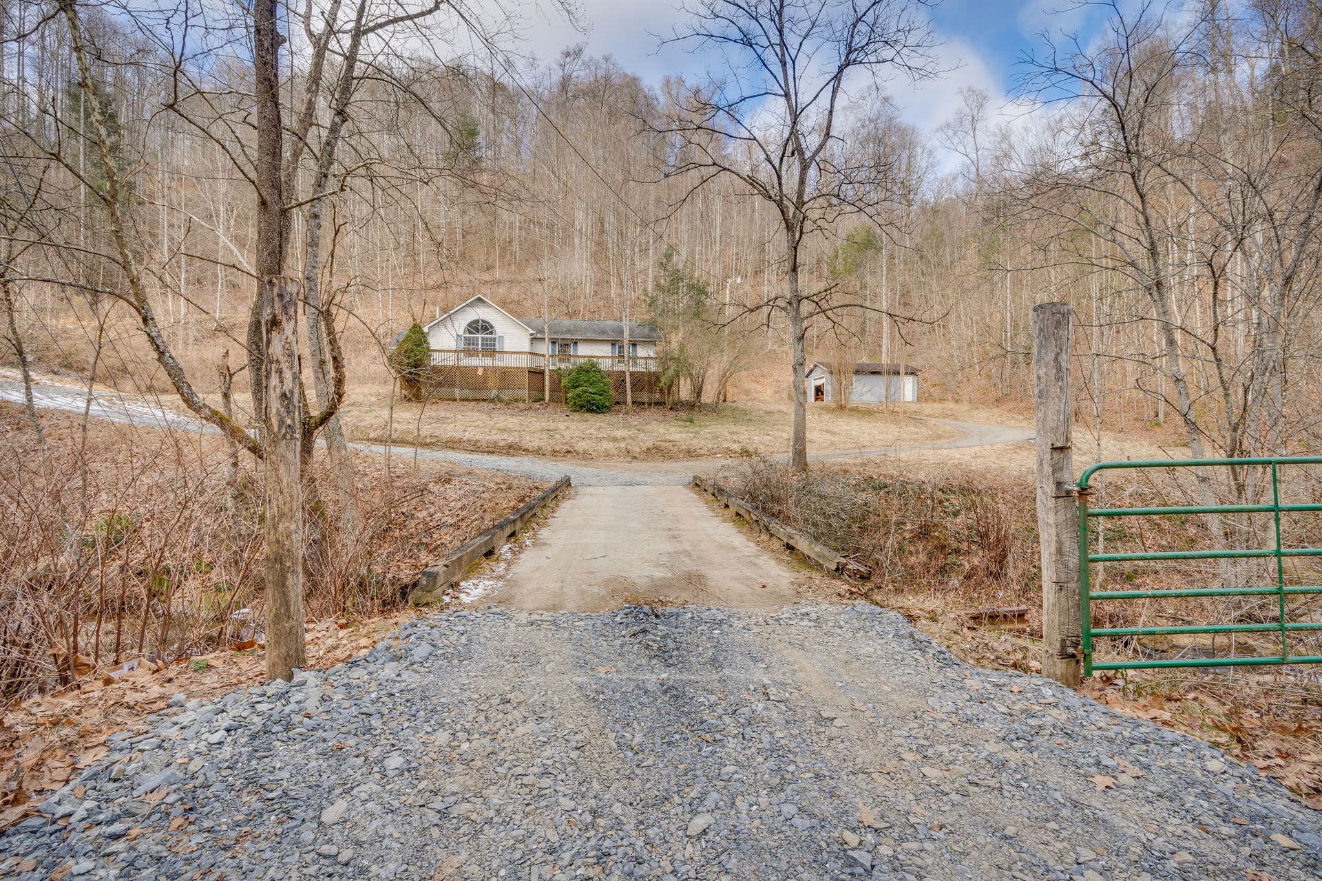 Gravel driveway leading to a small house nestled in a wooded area, with a green gate and bare trees.