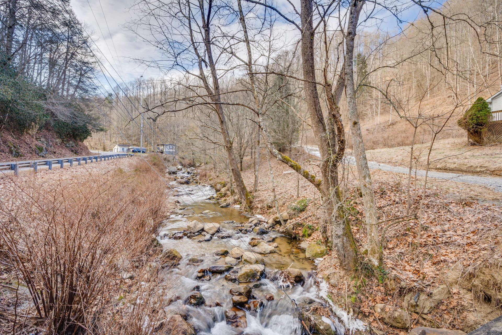 Creek flowing through a rocky bed, surrounded by bare trees, road, and dry brush.