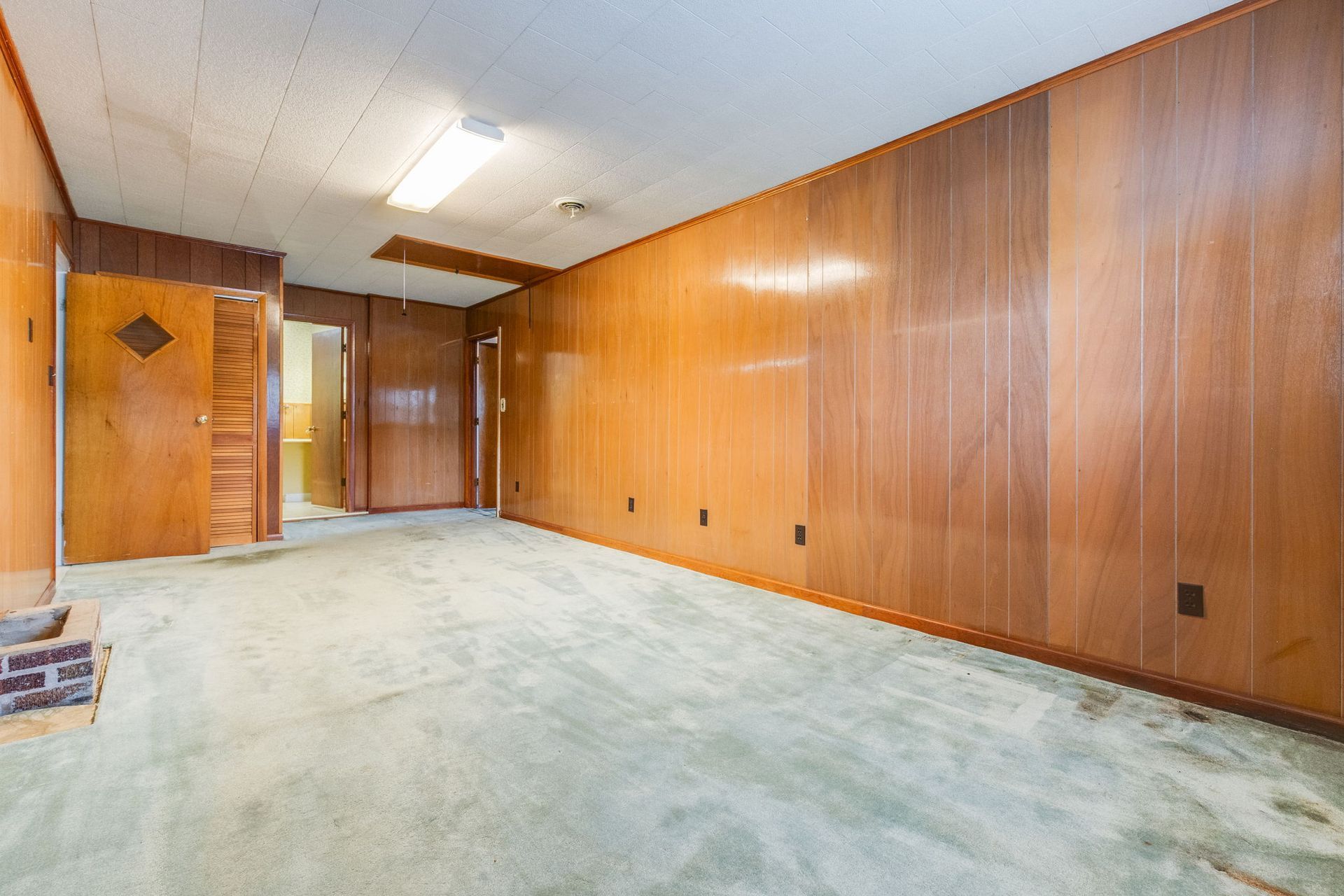 Empty wood-paneled room with green carpet, fluorescent ceiling lights, and a doorway at the far end