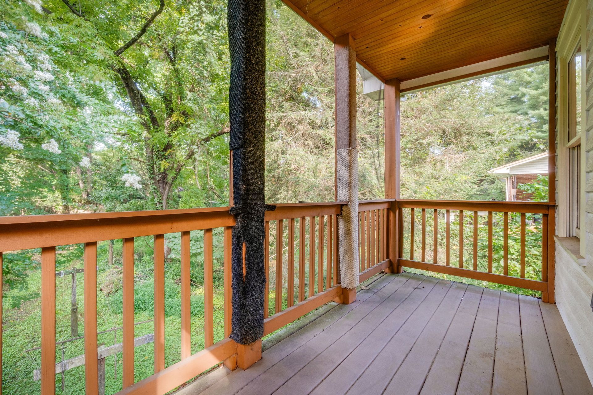 Wooden porch with railings and trees, featuring a covered ceiling and screened side opening.