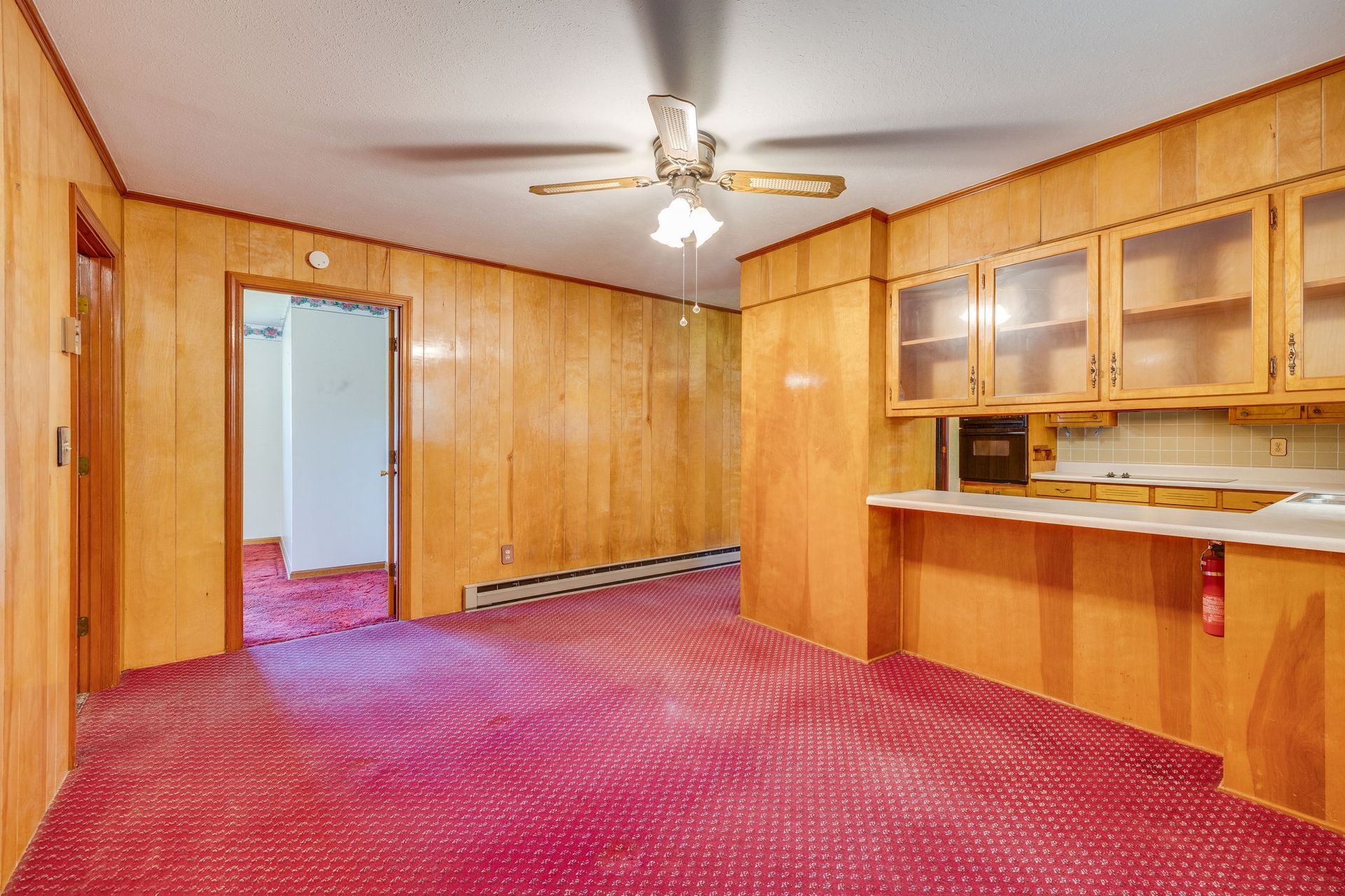 Empty room with wood-paneled walls, red carpet, ceiling fan, and built-in cabinets with a pass-through counter.