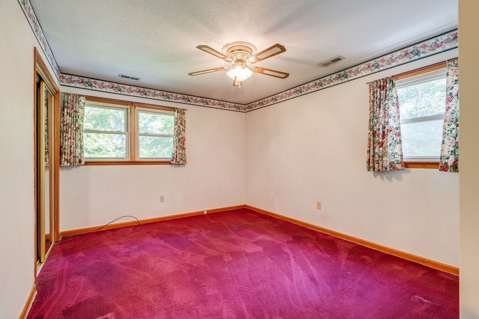Empty bedroom with magenta carpet, white walls, ceiling fan, and two windows with floral curtains