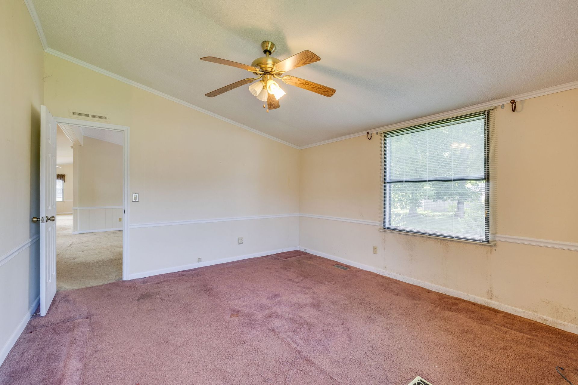 Empty beige bedroom with pink carpet, ceiling fan, and a window with blinds