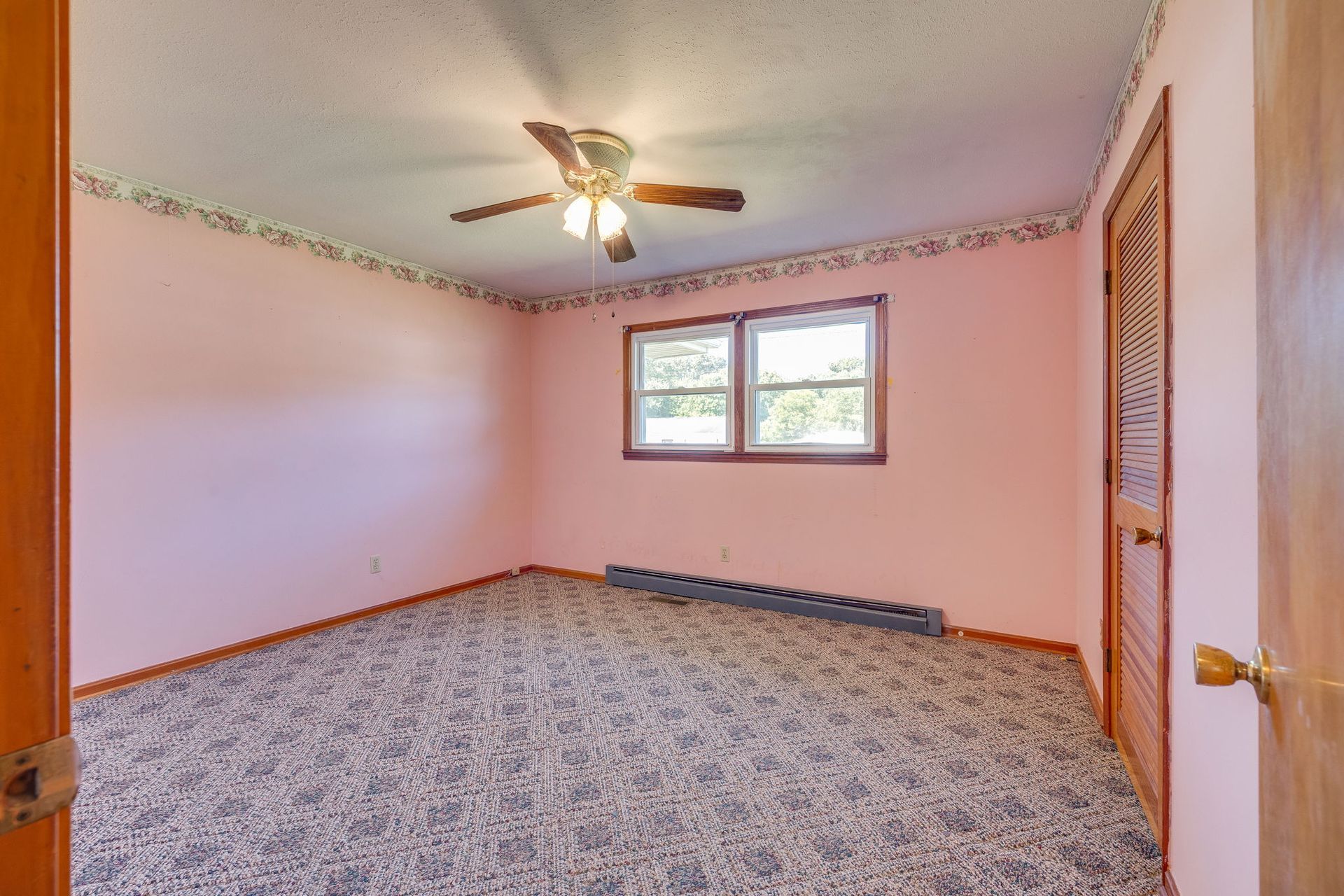 Empty pink bedroom with patterned carpet, ceiling fan, and a small window.