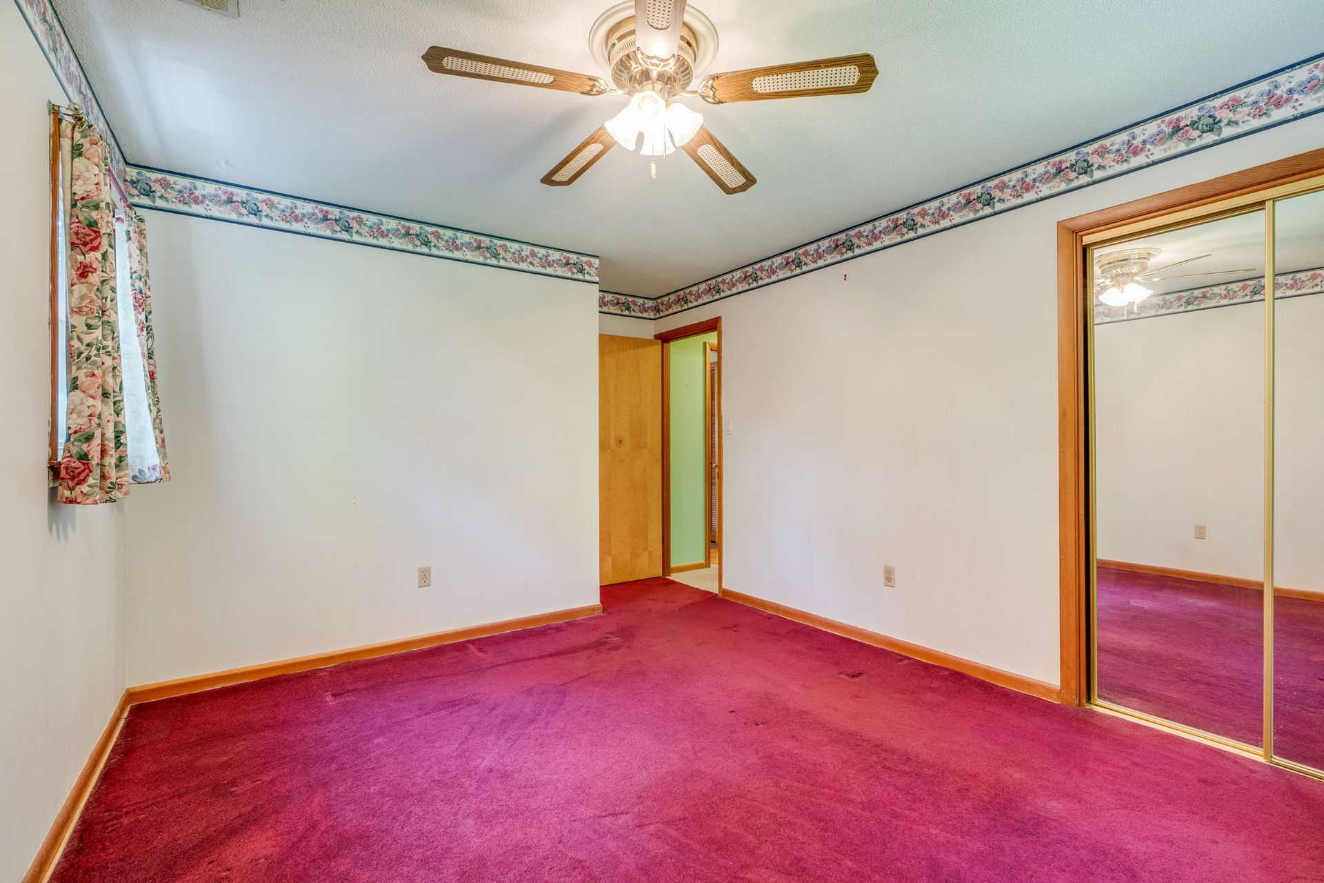 Empty bedroom with magenta carpet, white walls, mirrored closet, and ceiling fan light
