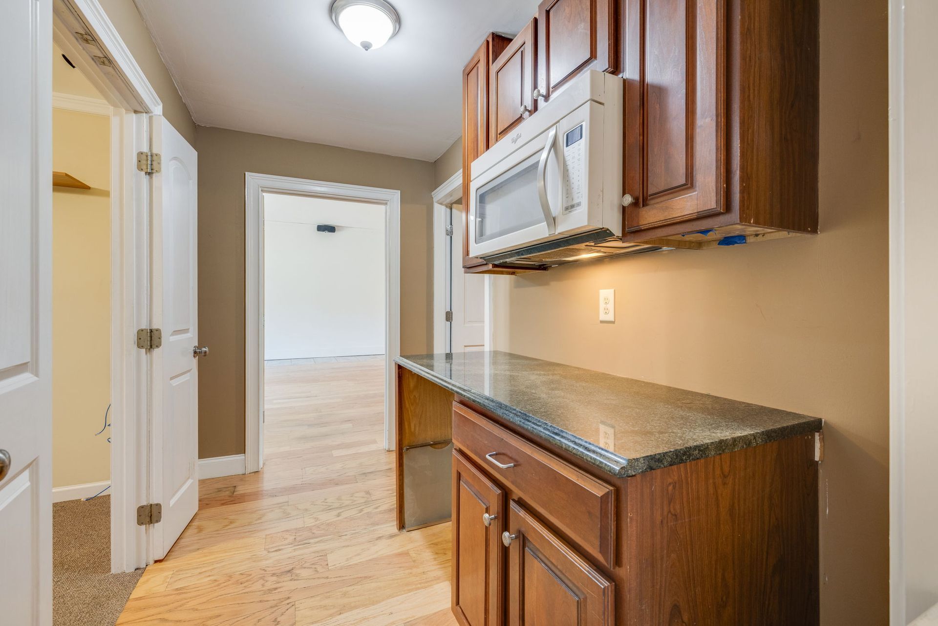 Narrow kitchen hallway with wood cabinets, granite counter, white microwave, and light wood floor