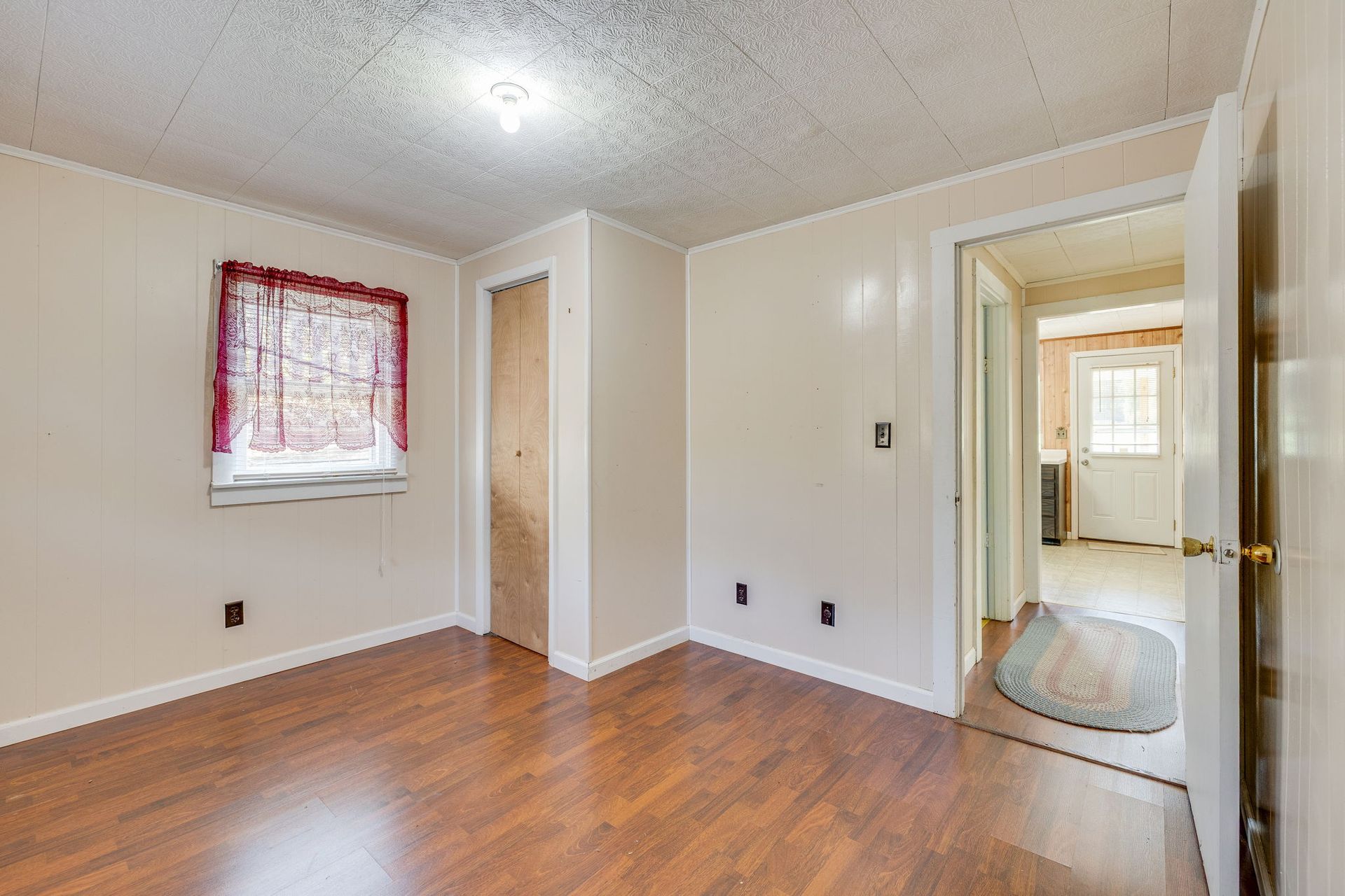 Empty beige bedroom with wood floors, small window with red curtain, and open doorway to a bright hall