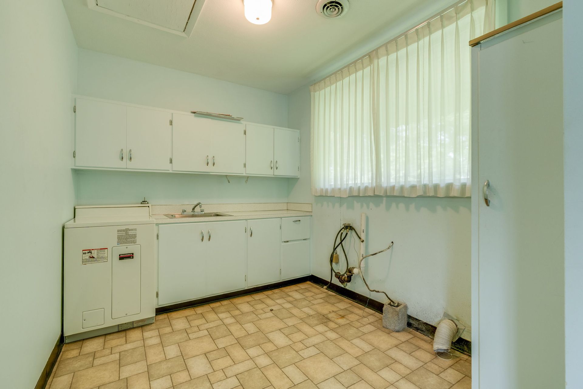 Empty white kitchen with cabinets, tiled floor, sink, and a large window with blinds.